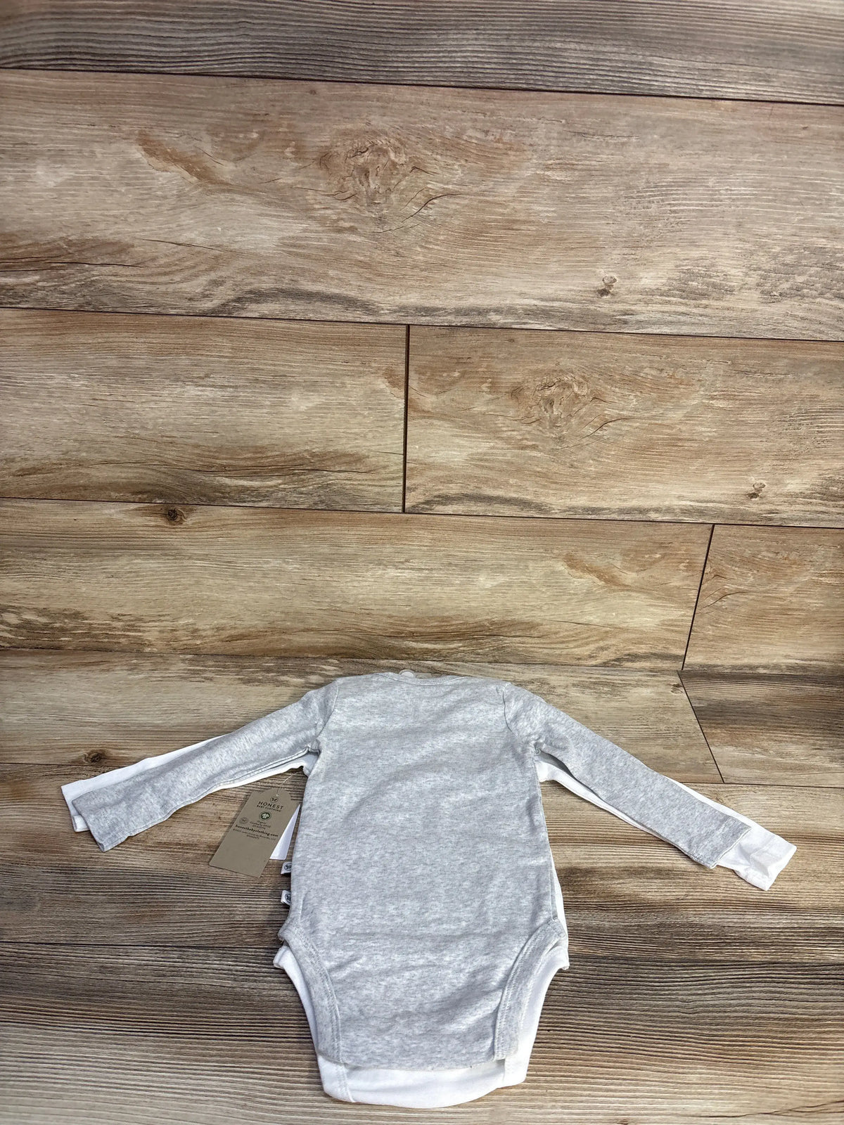 Front view of Honest Baby 6-9 month long sleeve bodysuit showcasing white organic cotton fabric on a wood plank background