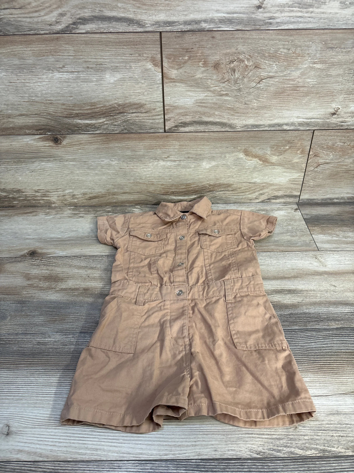Flat-lay of tan toddler utility romper with short sleeves, collar, button-front and chest and front pockets on wood background