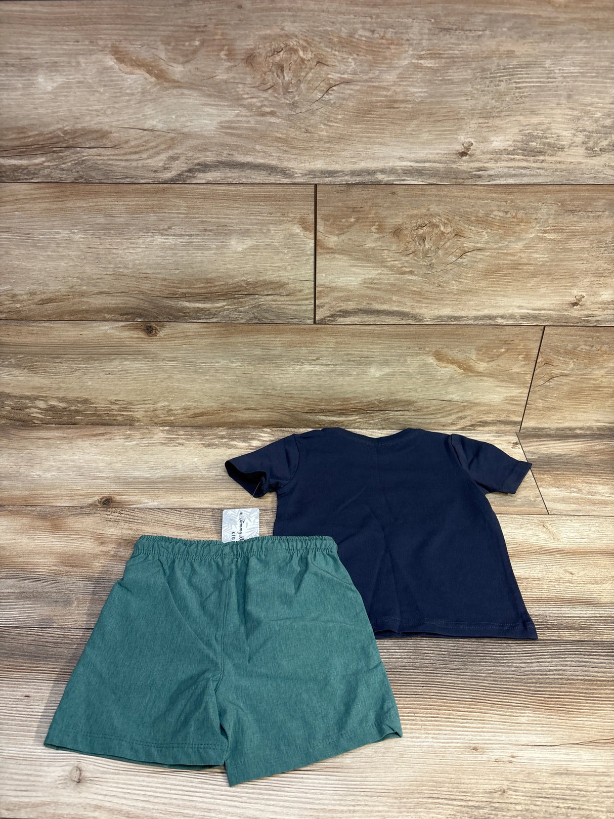 Back view of Tommy Bahama toddler navy dinosaur tee and teal board shorts with elastic waistband on wood background