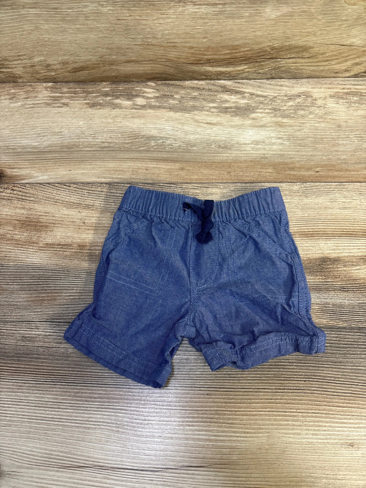 Front view of chambray baby shorts (3–6 months) showing elastic drawstring waistband, rolled cuffs, and front pockets on light wood background