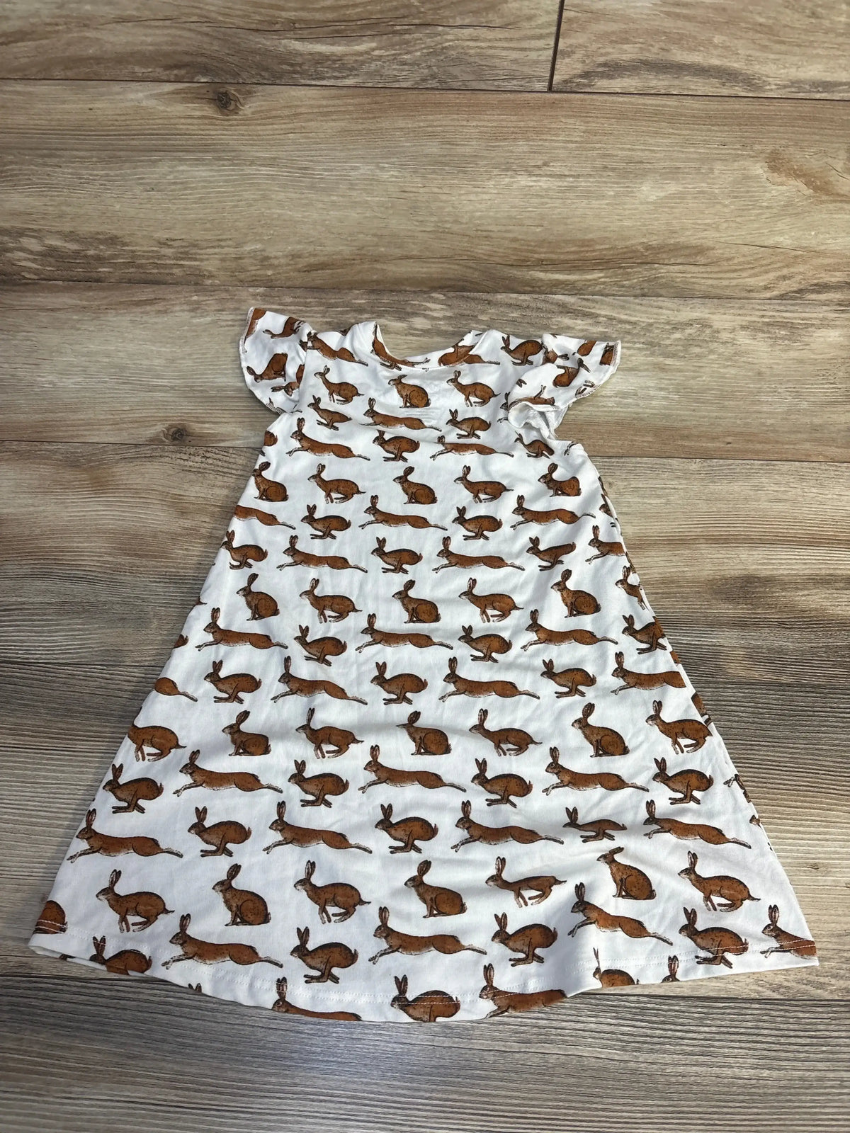 Laid-flat view of Milkmaid Goods Little Rabbit girls’ dress, size 2T, white with short sleeves and brown rabbit print on wood floor