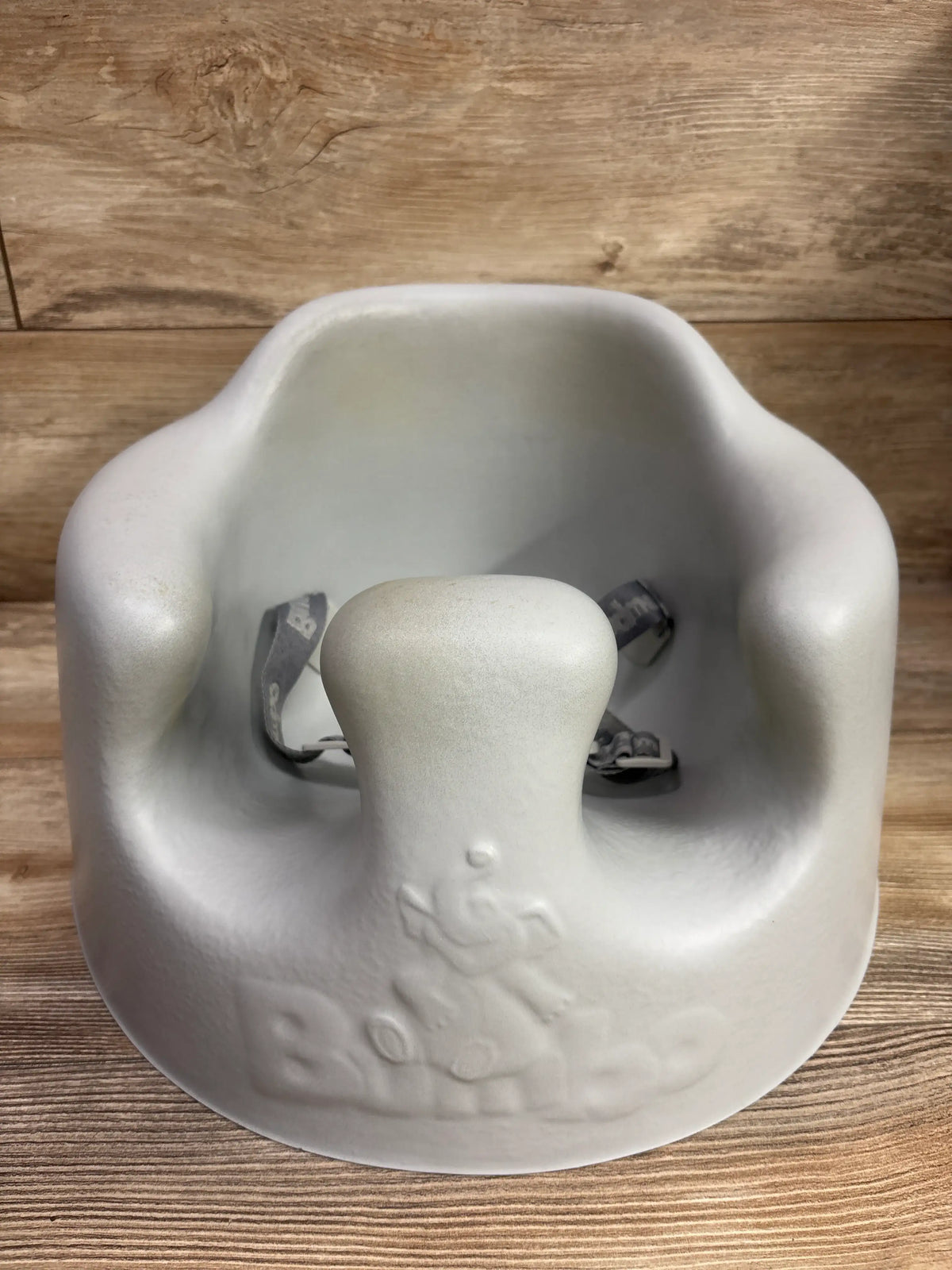 Front view of a grey Bumbo floor seat showcasing its molded seat and safety harness with a wood background