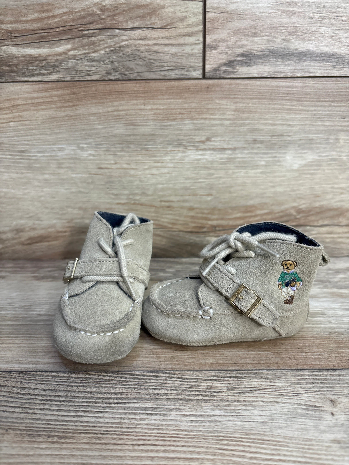 Beige suede baby shoes with buckle strap and embroidered teddy bear on wooden background