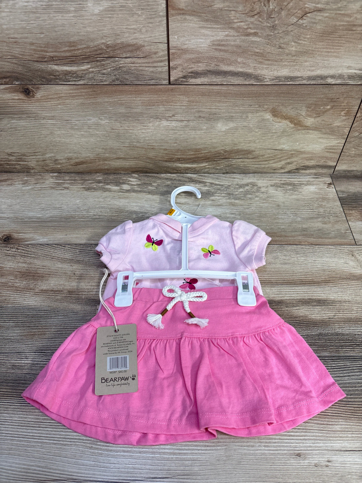 Front view of Bearpaw pink 2-piece polo and skort (6-9 months) with butterfly embroidery and braided drawstring on wood