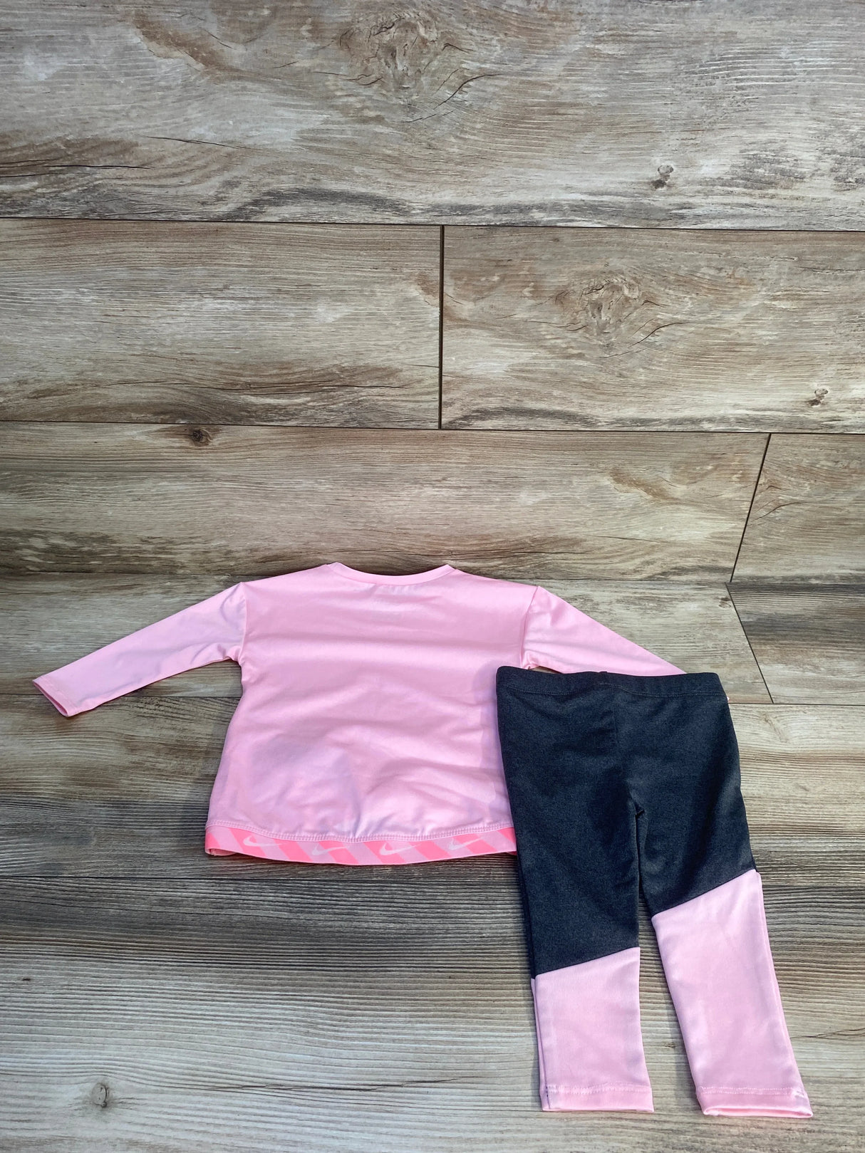 Flat-lay of Nike 2pc Dri-FIT shirt and leggings set in hot pink and charcoal, showing pink lower-leg panels on wood floor