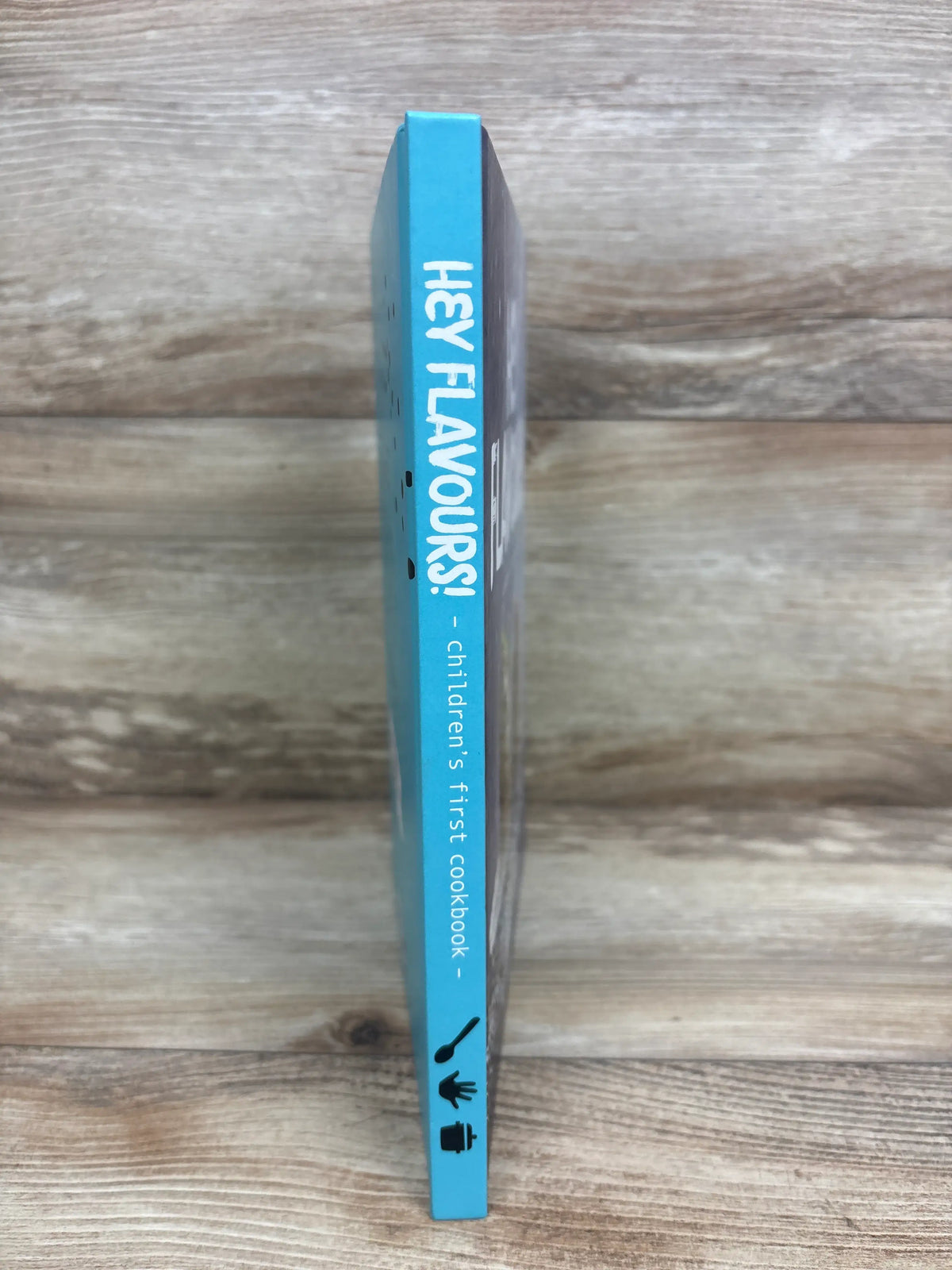 Side view of Hey Flavours! Children’s First Cookbook hardcover book showcasing turquoise spine with a wood-grain background