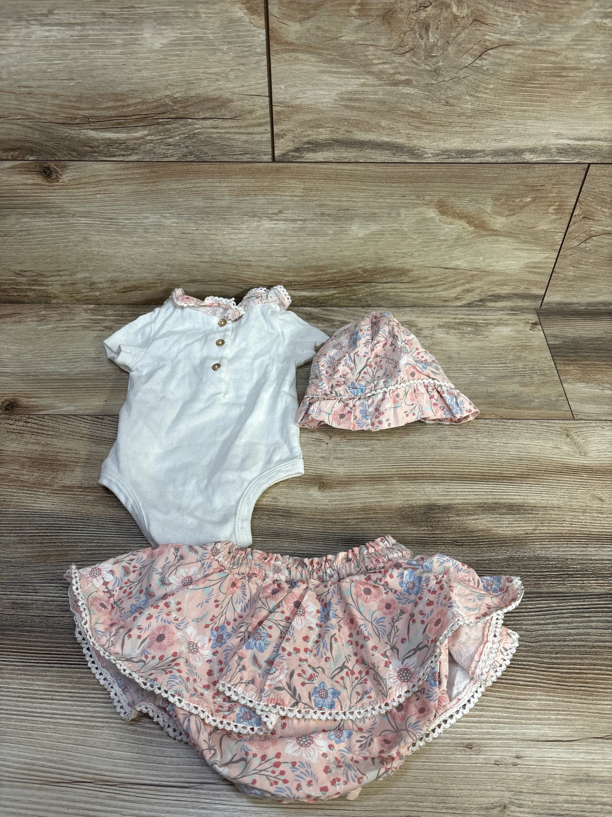 Flat lay of 3-piece baby bodysuit set, white bodysuit with lace collar, pink floral ruffle skirt and matching bonnet on wood
