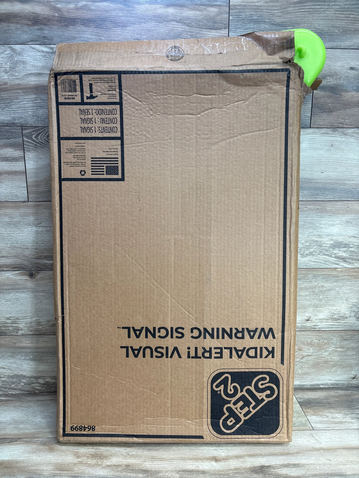 Cardboard box with Step2 kids' visual warning sign packaging on wood floor, partially opened with green plastic visible