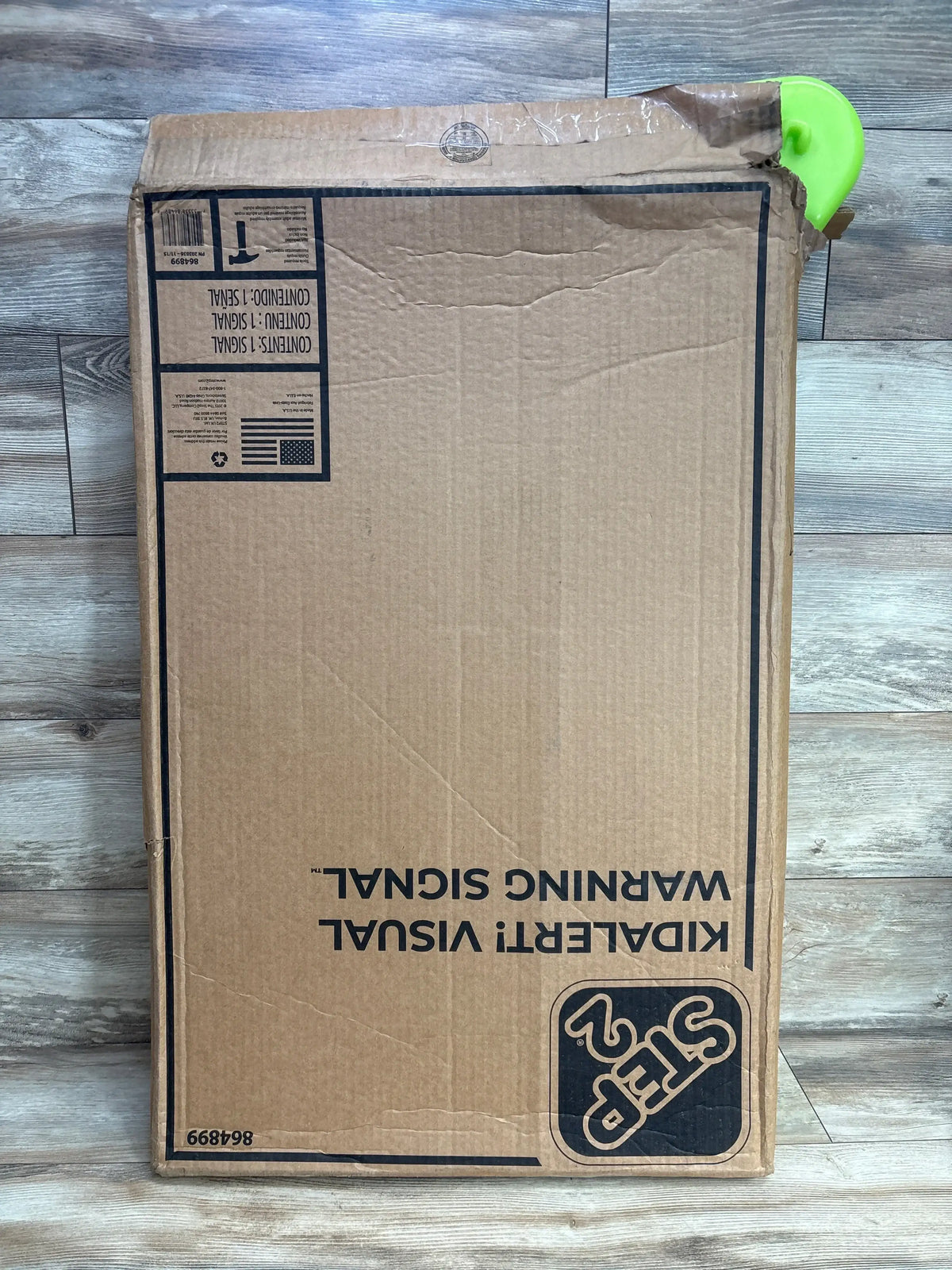 Cardboard box with Step2 kids' visual warning sign packaging on wood floor, partially opened with green plastic visible