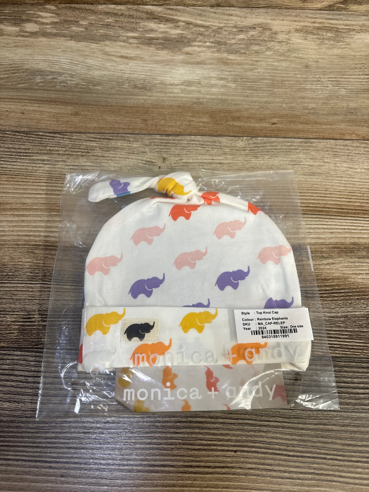 Top view of a Monica + Andy white top knot cap showcasing a rainbow elephant pattern in plastic packaging on a wood surface