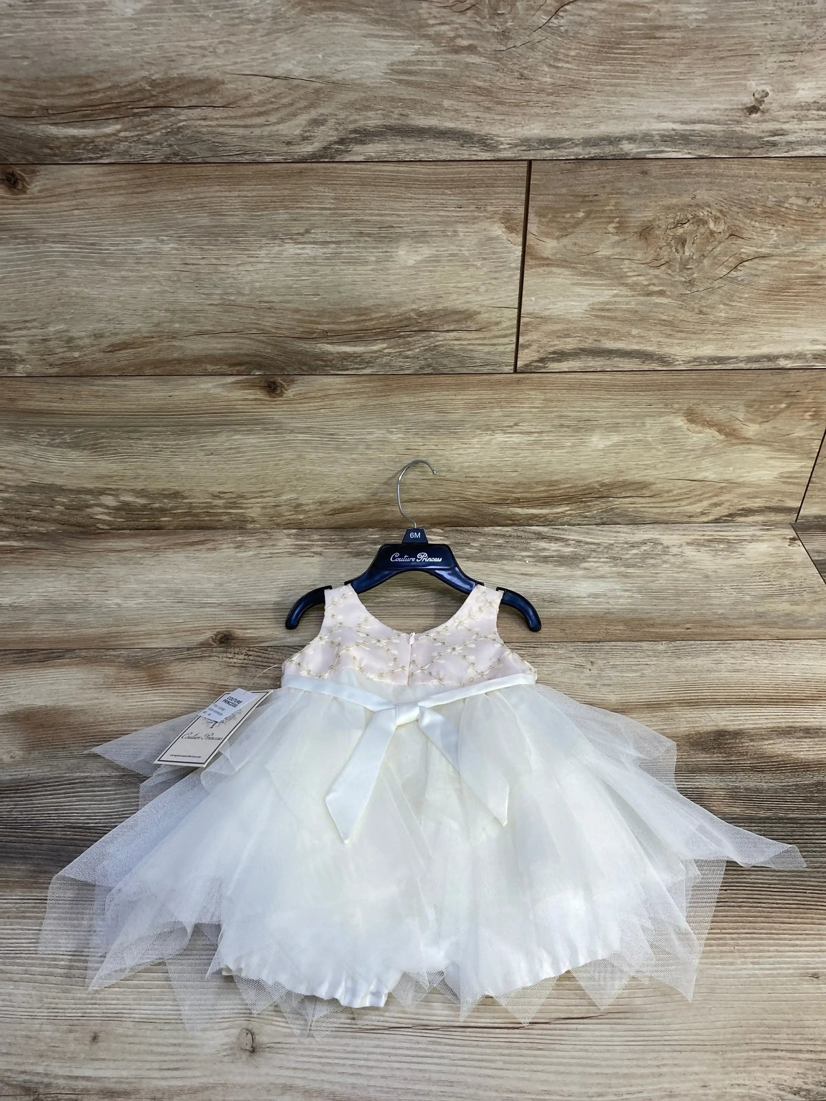 Front view of pink embroidered pearl dress and bloomers with bow and layered tulle skirt on a hanger against wood background