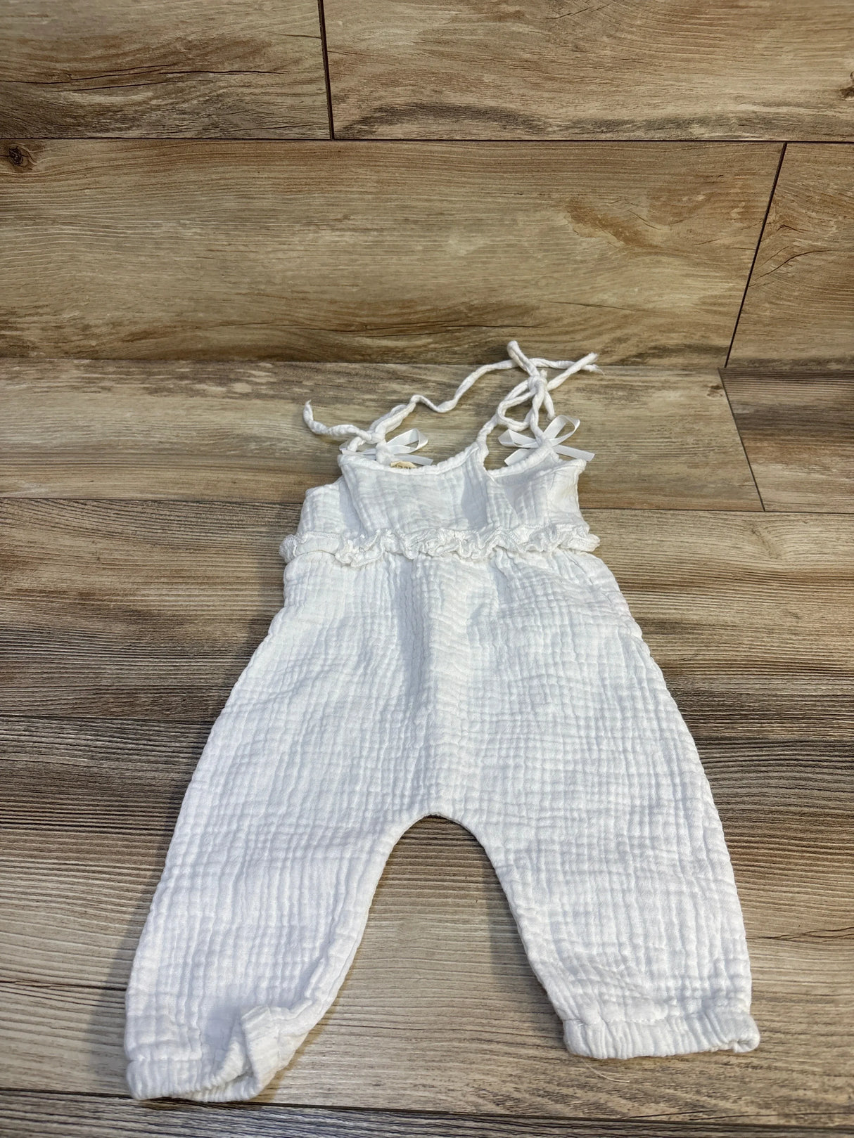 Flat lay of white muslin tank jumpsuit, size 6-9 months, showing tie shoulder straps, ruffle waist and gathered leg cuffs on wood floor