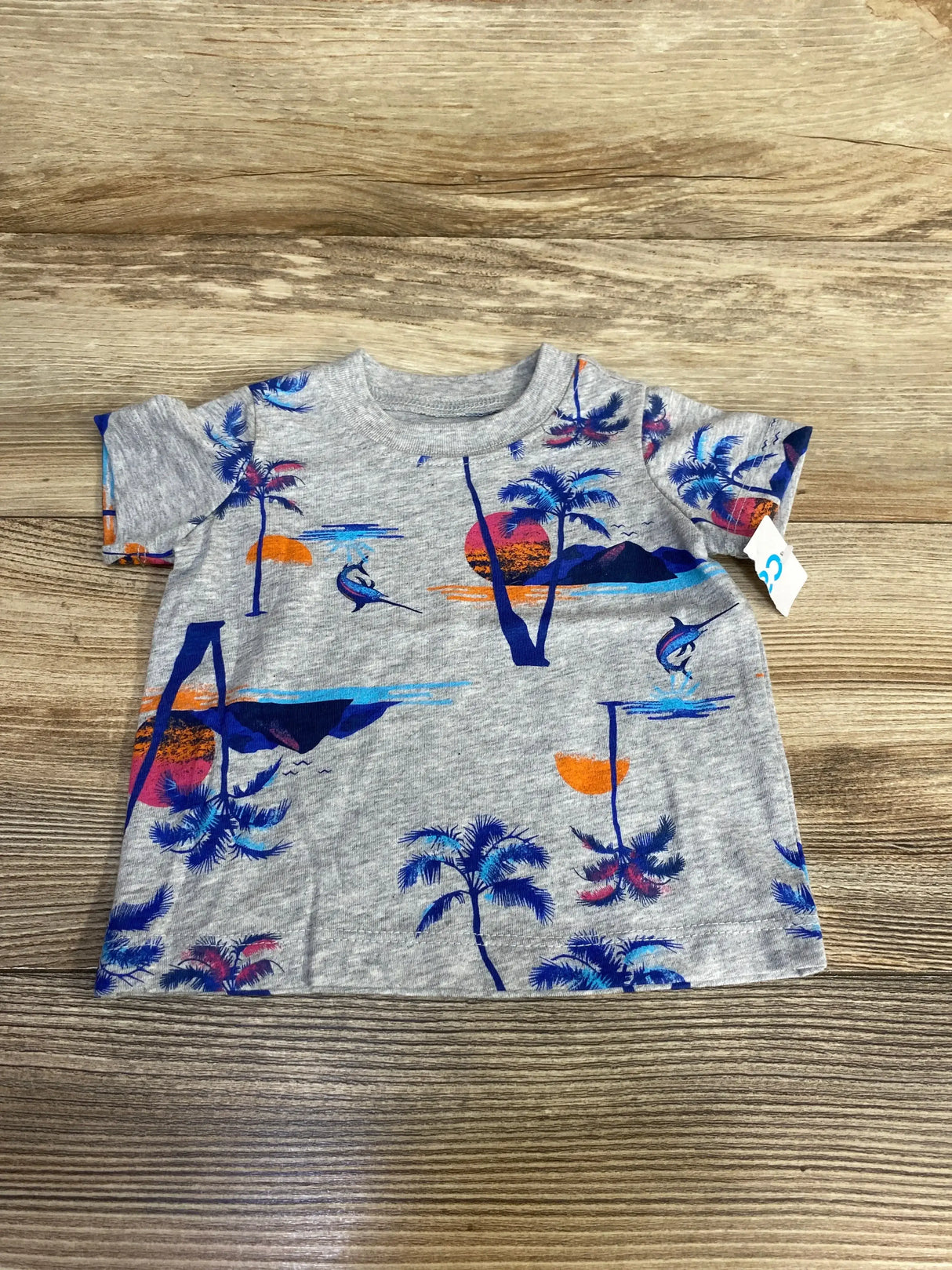 Flat lay front view of grey Carter’s short sleeve baby shirt showcasing orange and blue sunset, palm tree and marlin print on wood background