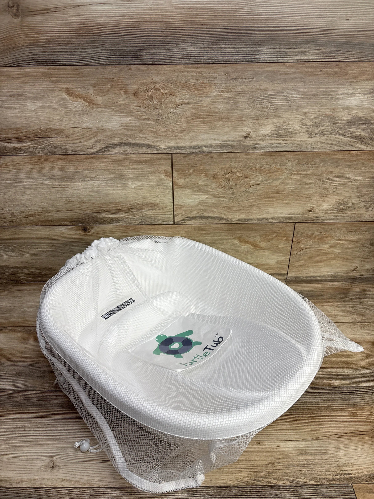 Angled top-down view of TurtleTub Swaddle Bath Tub showing white mesh fabric, contoured seat and TurtleTub logo on wood floor