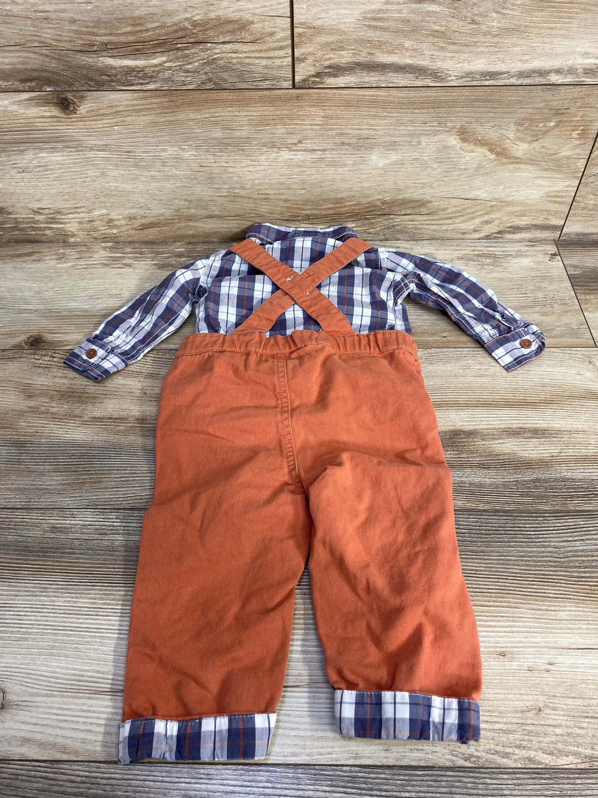 Toddler outfit with long-sleeve plaid shirt and burnt orange suspenders pants on wooden floor