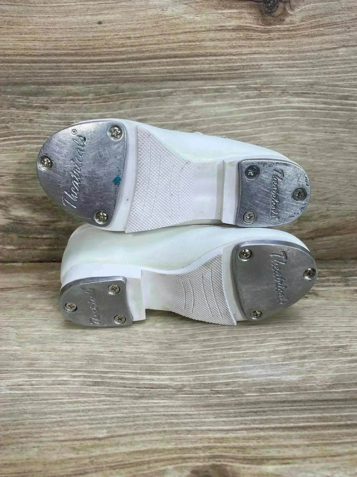 Bottom view of child white tap buckle shoes size 6.5C showing metal taps screwed to heel and toe with wood floor background