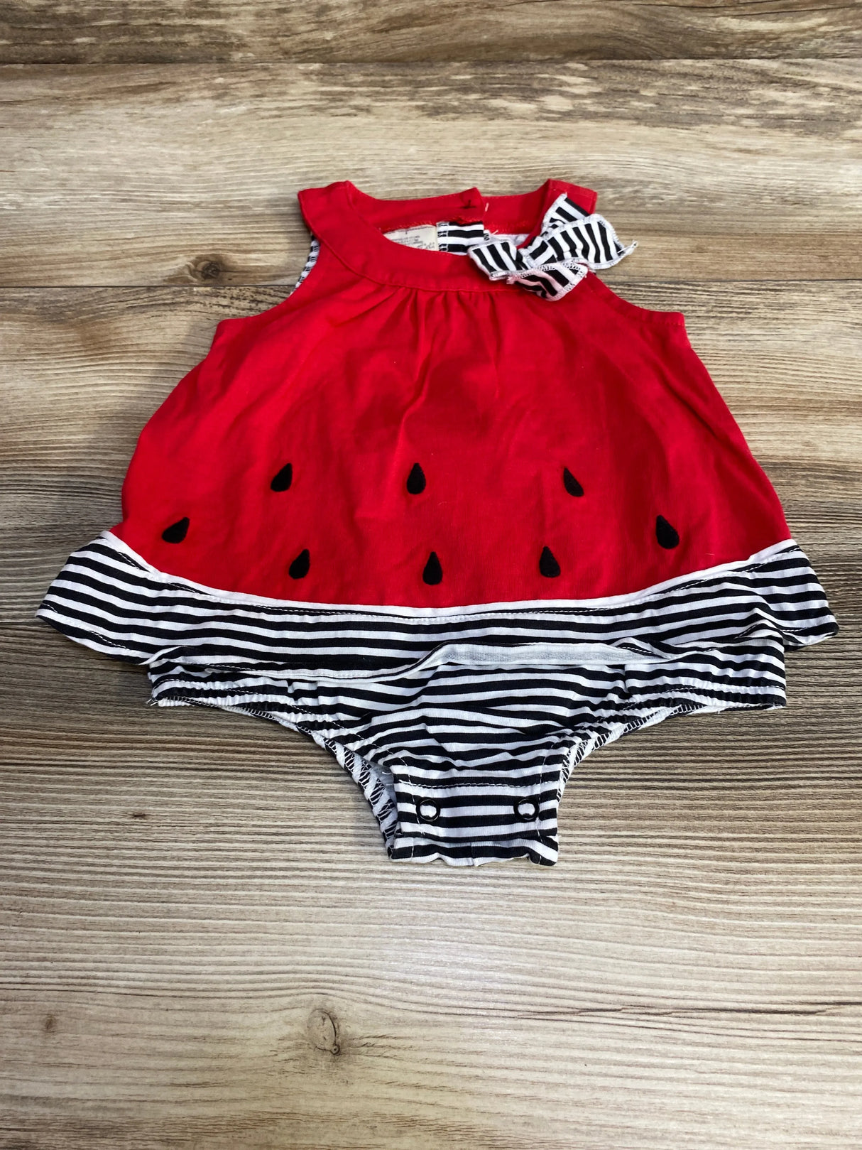 Flat lay of red watermelon skirted sunsuit, 3-6m, showcasing black seed appliques and black-and-white striped bloomers on wood