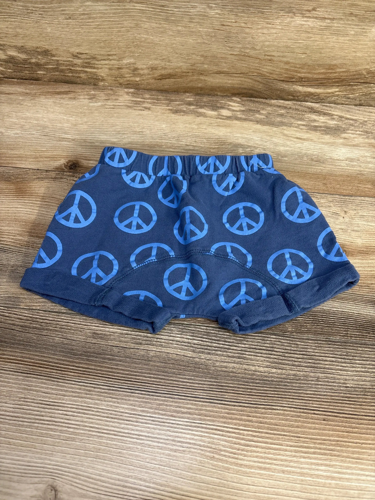 Front flat lay of navy Cotton On baby shorts showcasing blue peace sign print and elastic waistband on light wood background