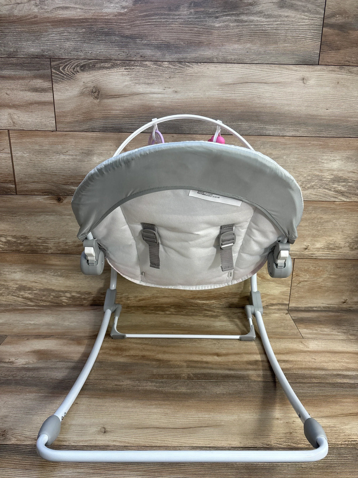 Rear view of Bright Starts Rosy Rainbow rocker with gray seat back, harness straps, toy bar, white frame on wood floor