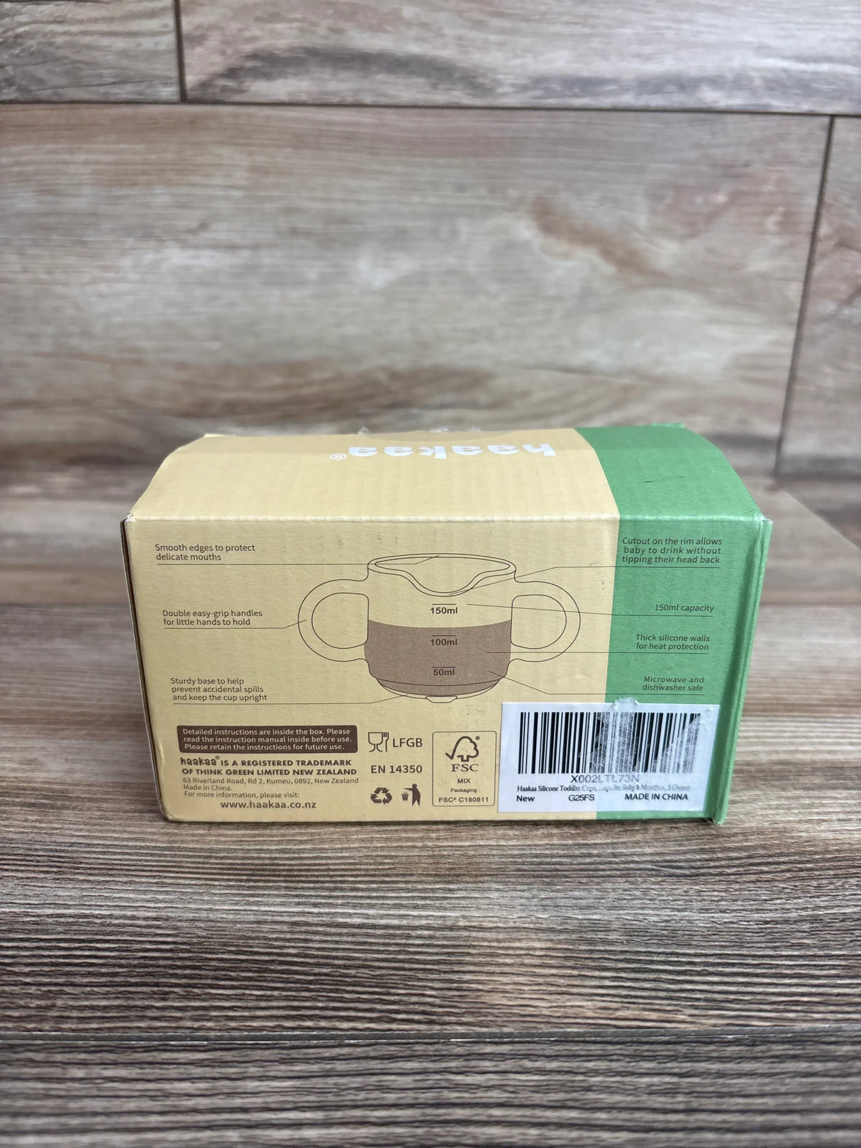 Back view of Haakaa silicone baby drinking cup box showing cup illustration with 50/100/150 ml markings and dual handles on wooden tabletop