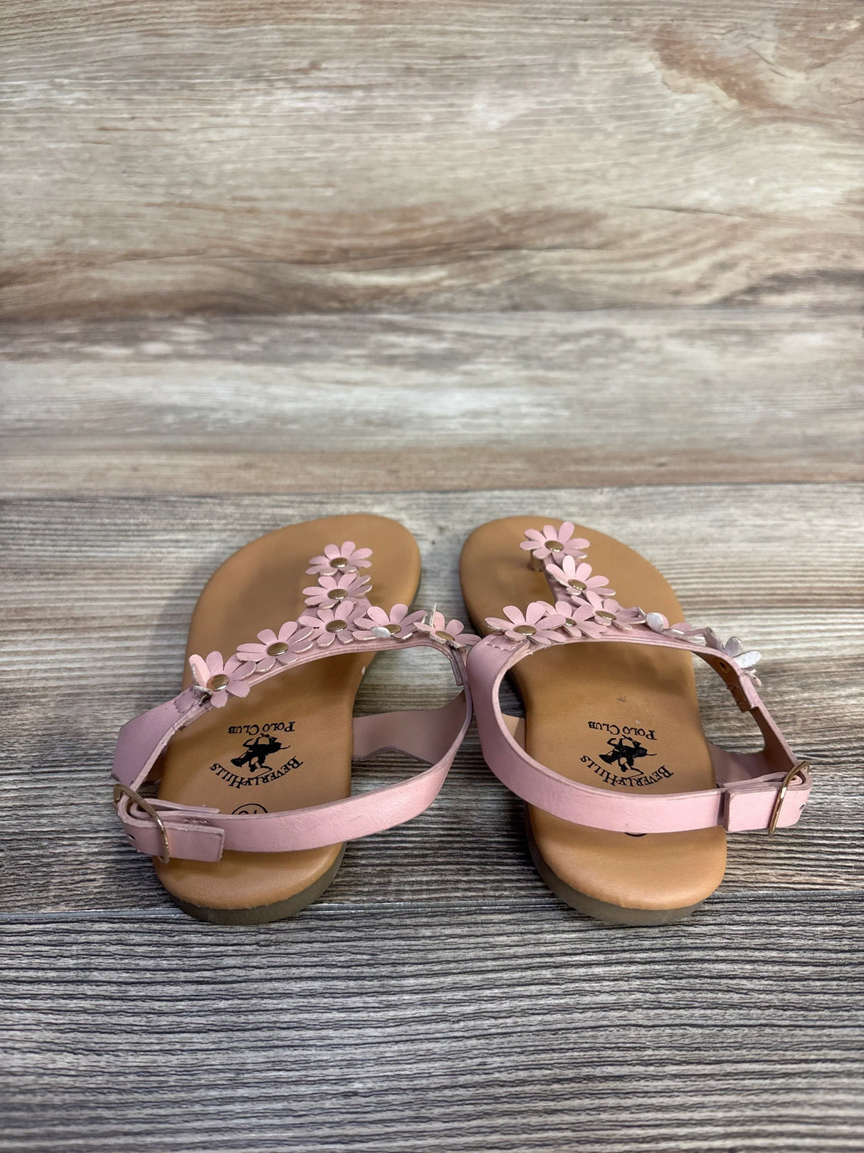 Top-down view of pink Beverly Hills Polo Club toddler sandals showing daisy floral strap accents and ankle buckles on gray woodgrain background