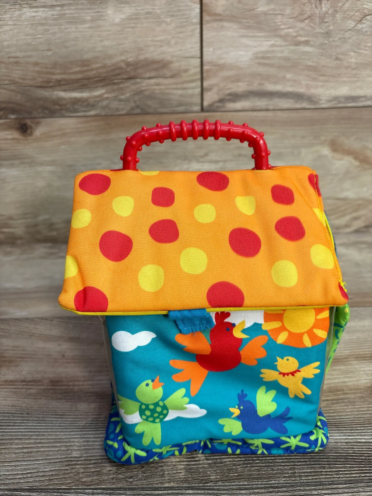 Front view of Manhattan Toys Put & Peek Birdhouse showing orange polka-dot roof, red textured handle, and colorful bird print on blue base on wood floor