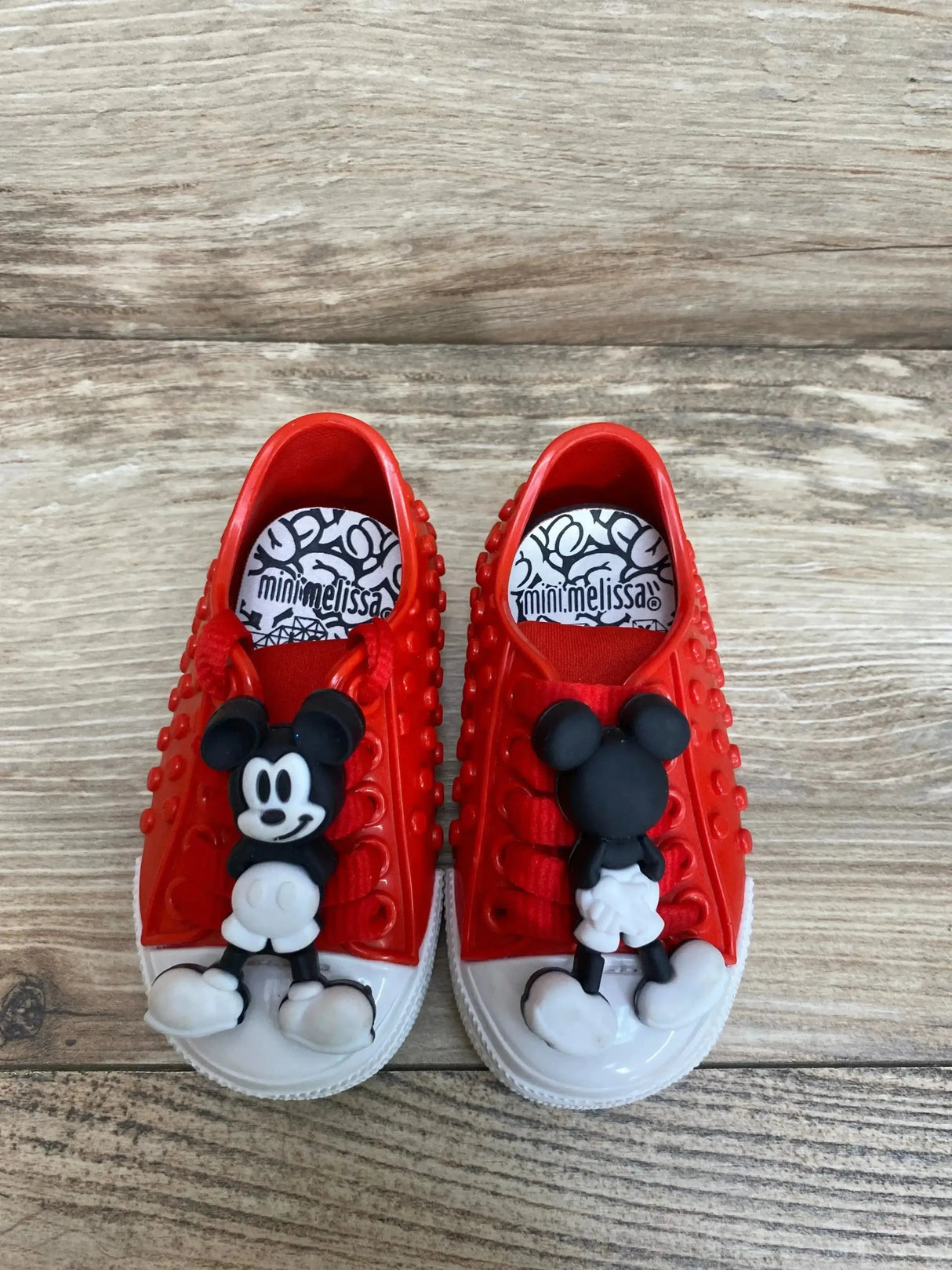 Top view of red Mini Melissa Polibolha x Disney sneakers with Mickey Mouse charms, white toecaps and studded sides on wood