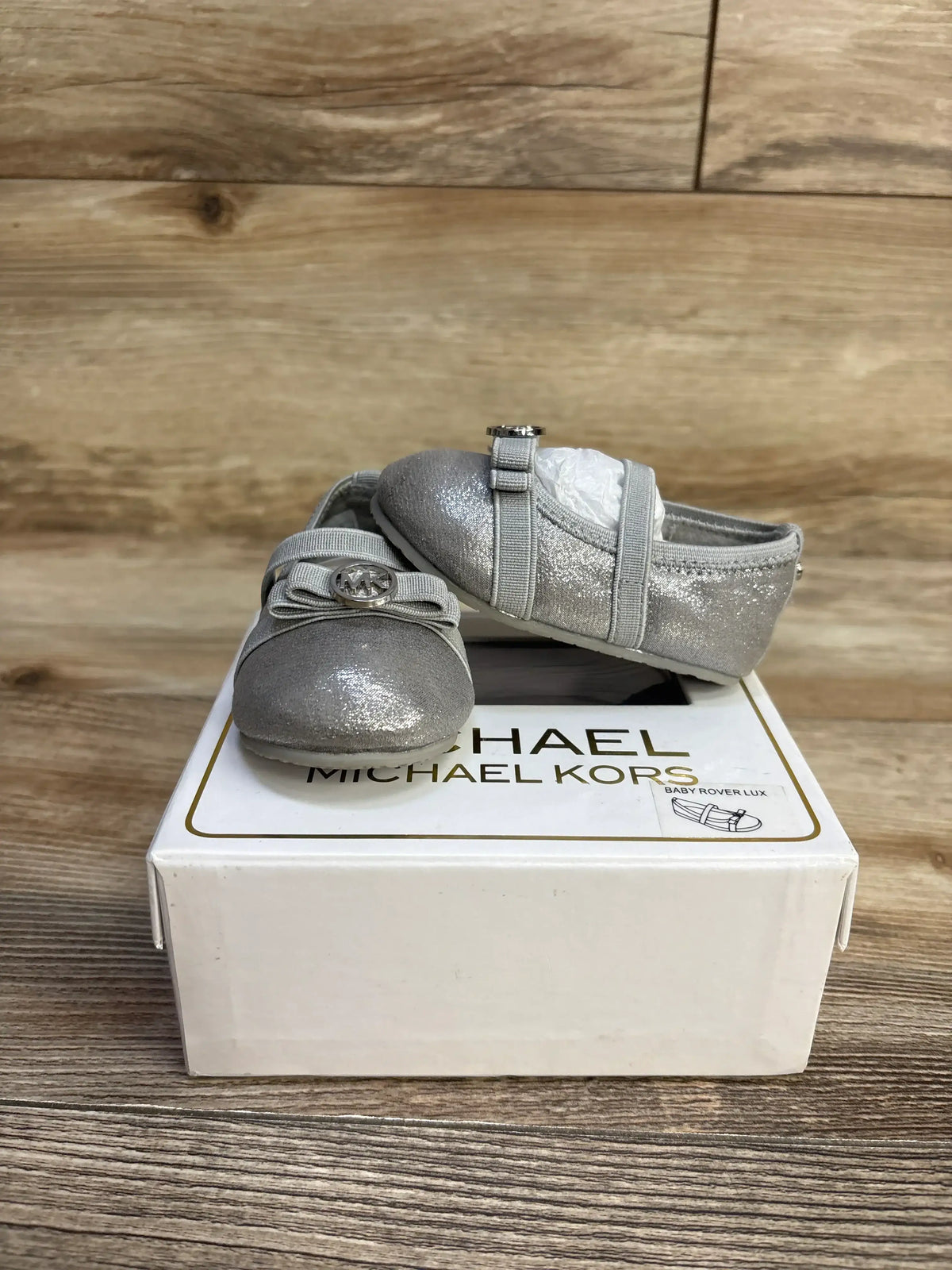 Front and side view of Michael Kors Baby Rover Lux silver shoes with glitter finish and MK bow on white box on wood floor