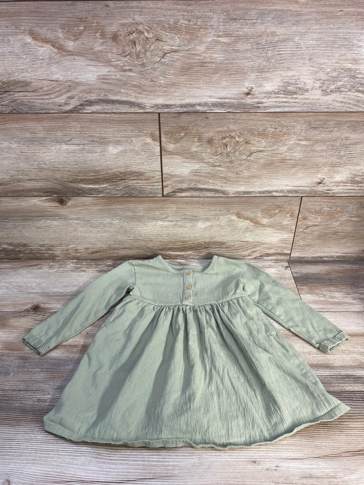 Flat lay of Cat & Jack long-sleeve henley dress (3T) showing sage green texture, wooden buttons and gathered skirt on wood-plank background