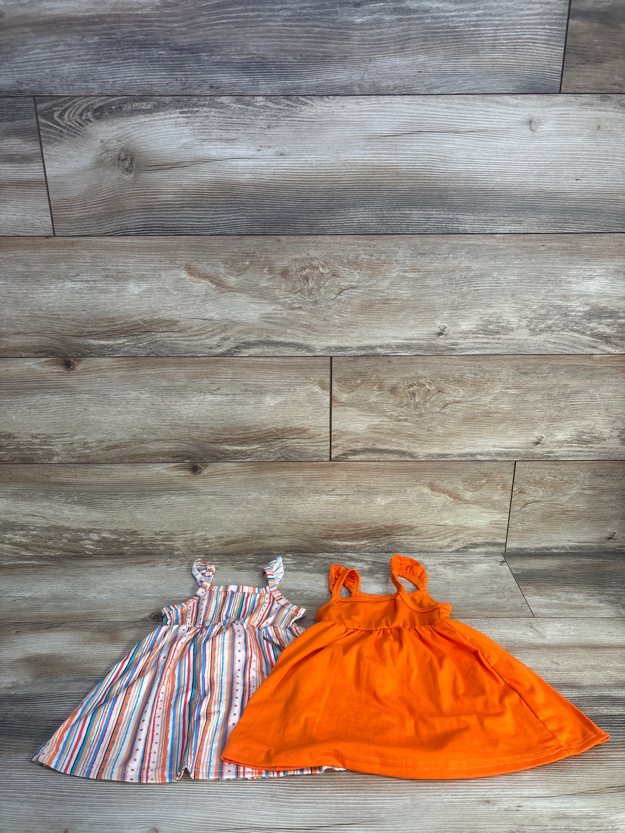 Flat lay of two baby sleeveless dresses showing a solid bright orange dress and a multicolor striped dress on wood background