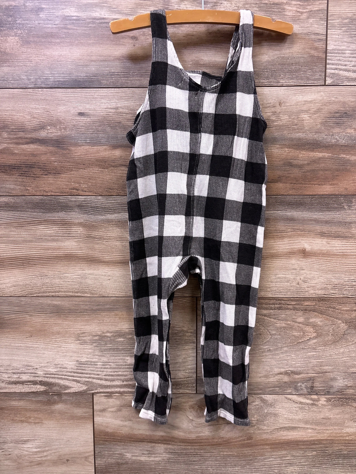 Black and white plaid sleeveless baby jumpsuit hanging on wooden hanger against wood panel background