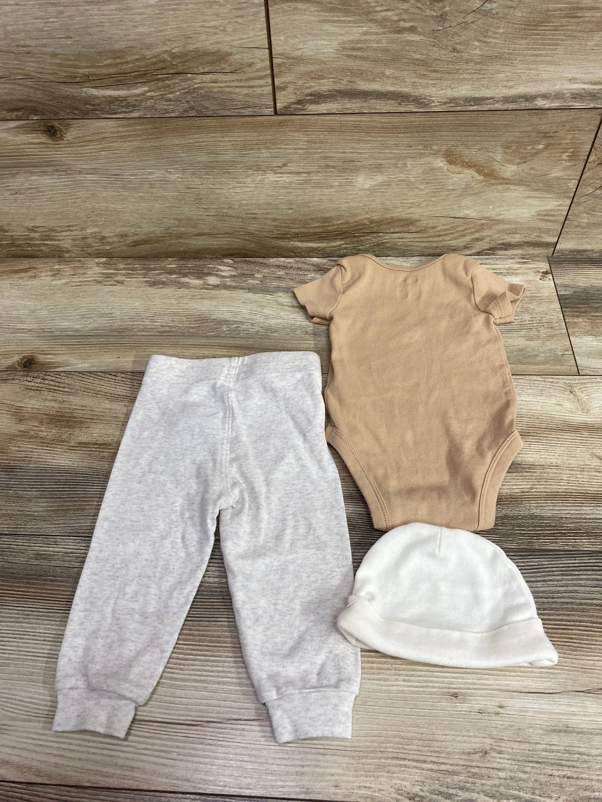 Nike 3-piece baby set featuring a tan bodysuit, grey pants, and a white hat on a wood floor.