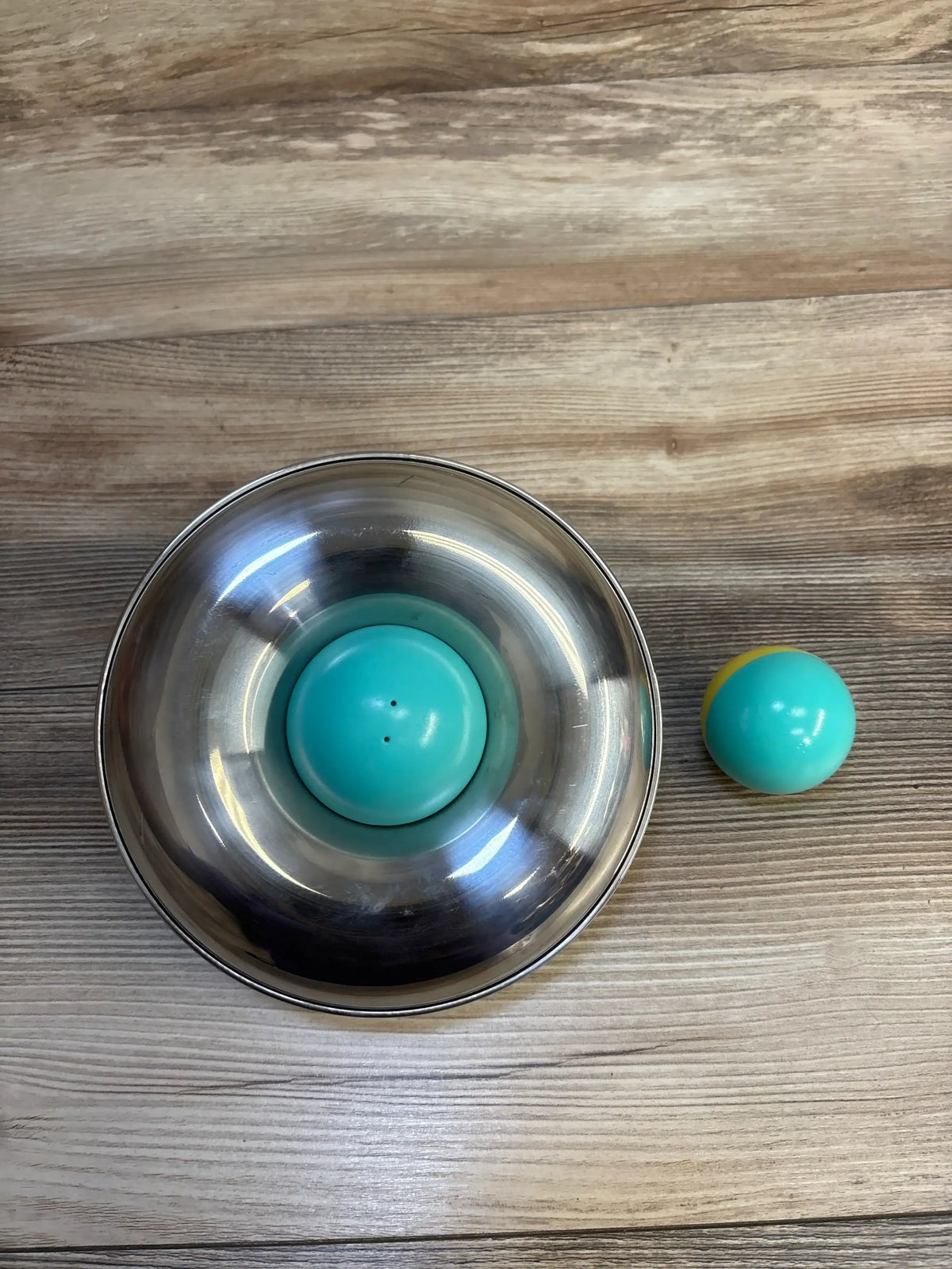 Top-down of Lovevery Stainless Steel Tip and Turn, stainless bowl with turquoise spinner and teal ball on wood