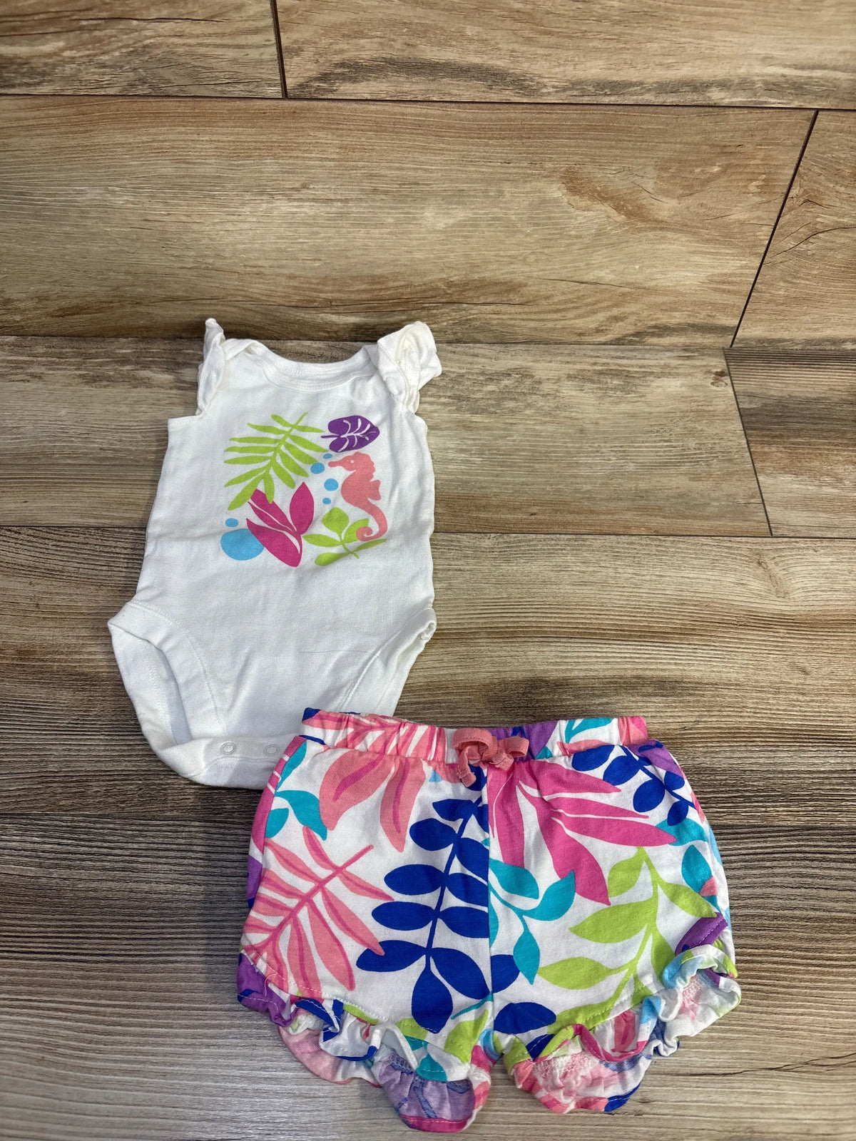 Flat lay of Children’s Place white bodysuit and colorful printed shorts set on wood flooring