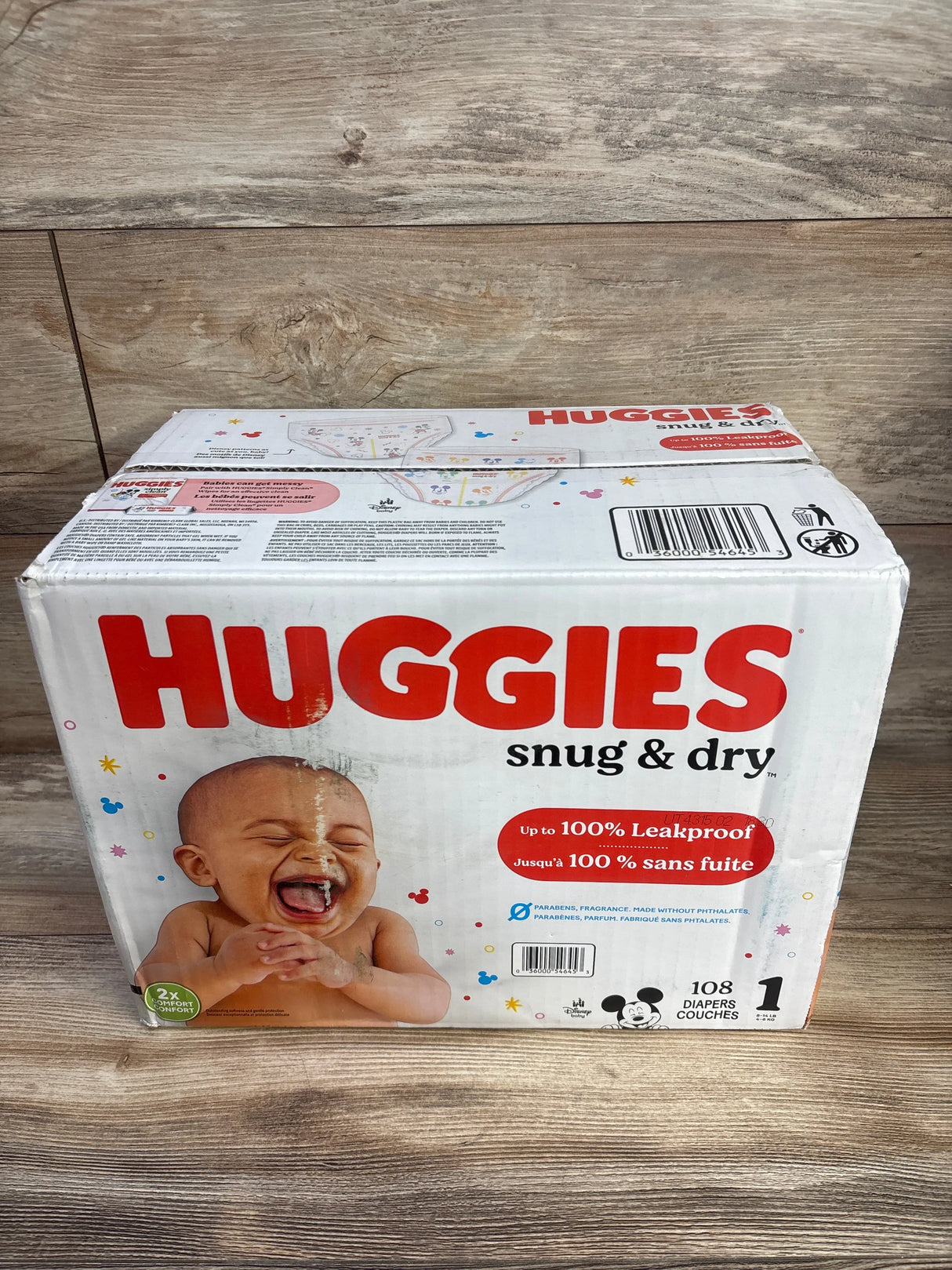 Front of Huggies Snug and Dry size 1 box, 108 diapers, baby photo and leakproof claim on wood surface