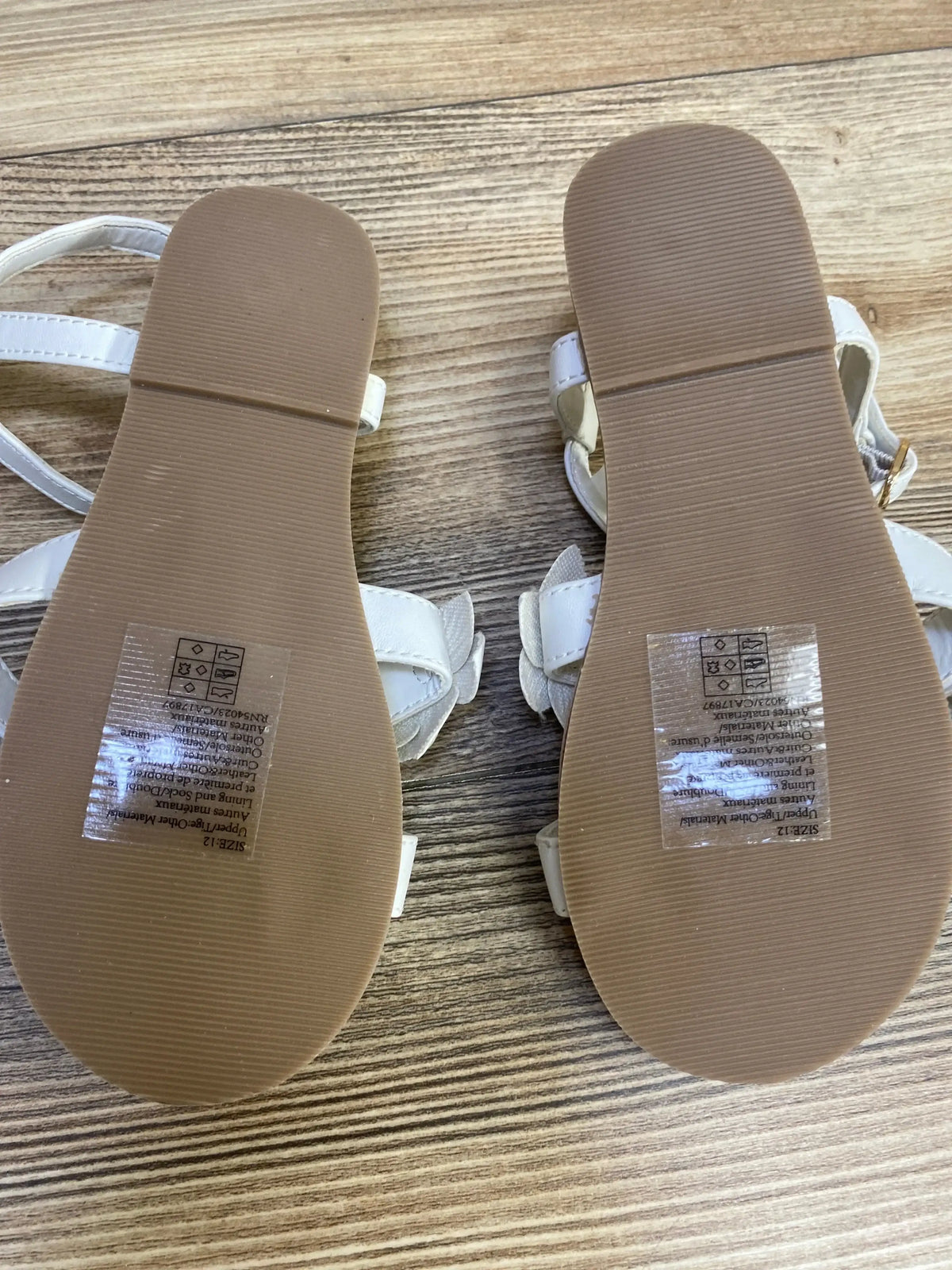 Sole view of Janie and Jack flower sandals white size 12c showcasing tan rubber soles with brand stickers on wood plank floor