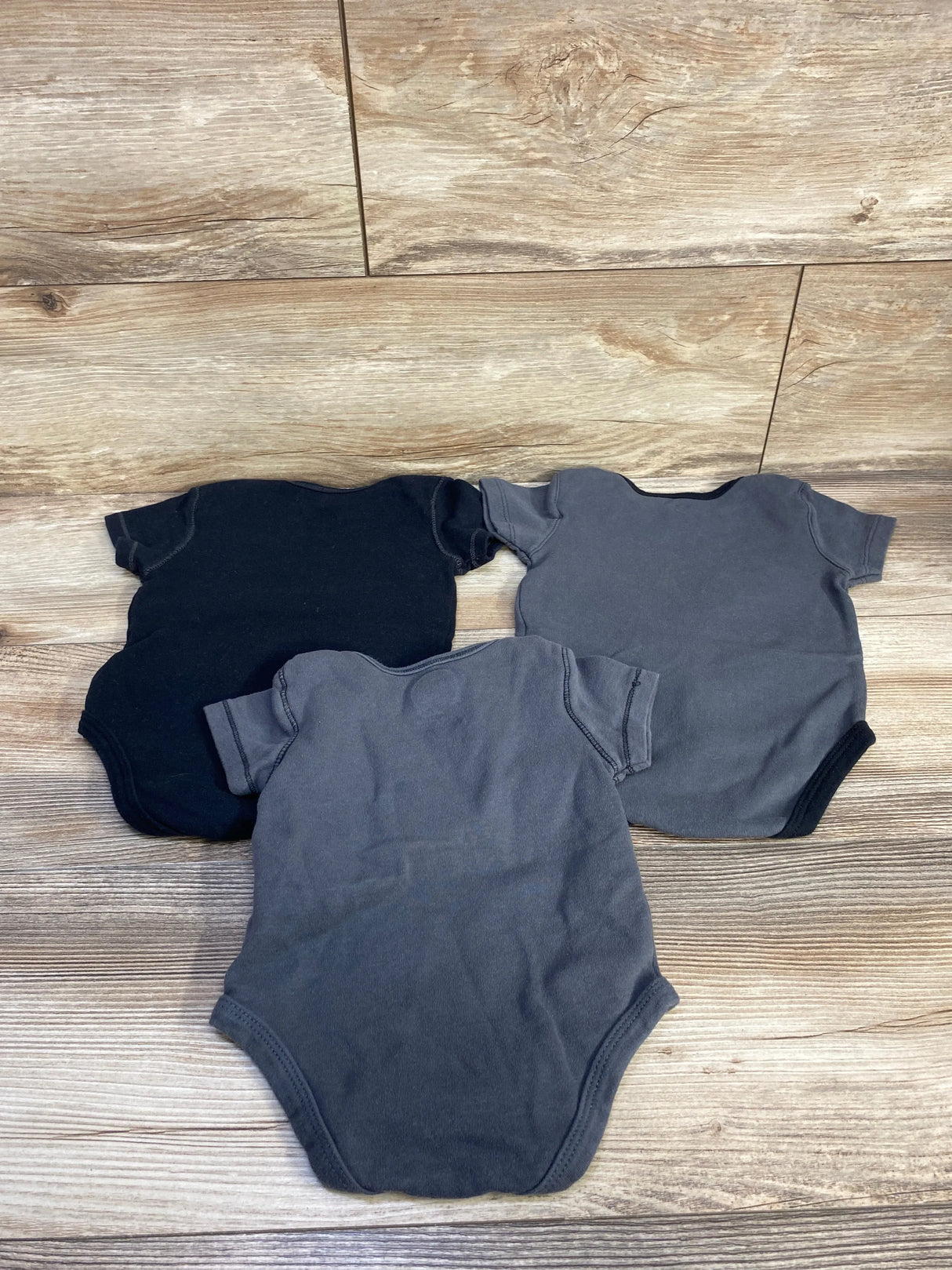 Top-down view of 3-pack baby bodysuits showcasing short sleeves and crew necks in dark grey and black on wood background