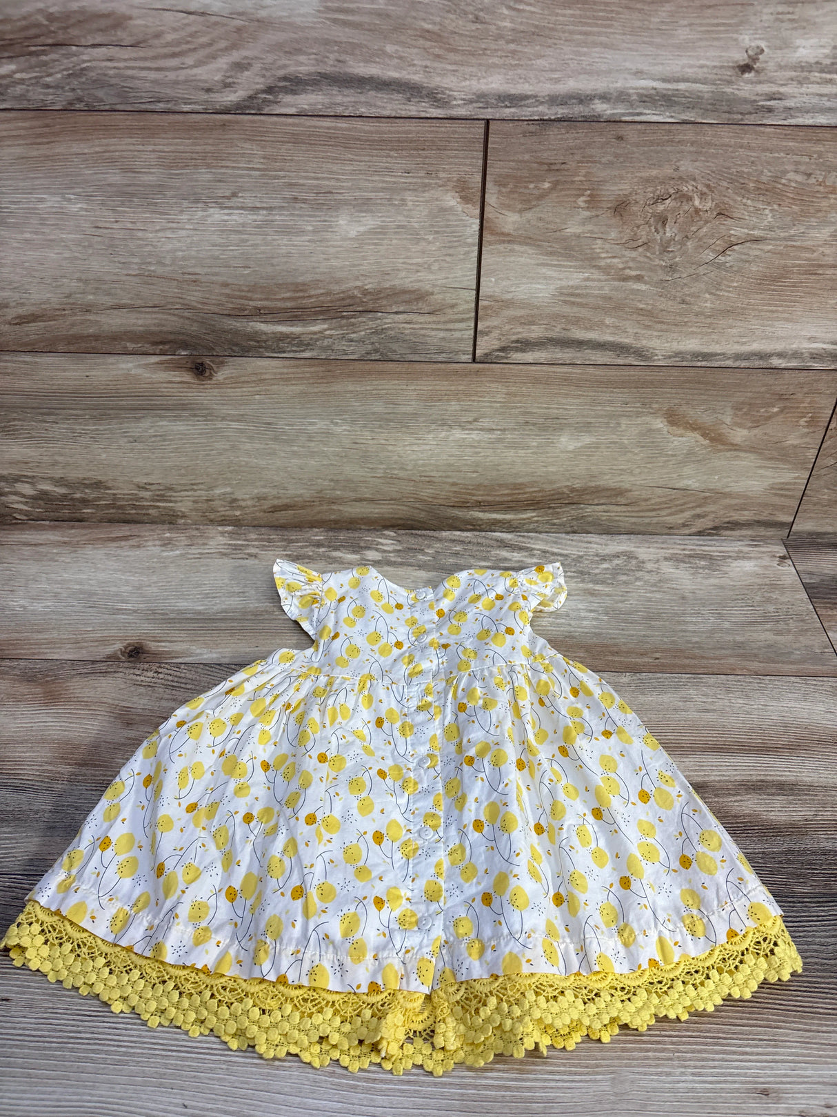 Flat lay of Du Pareil Au Meme baby lemon-print dress showcasing flutter sleeves and yellow crocheted lace hem on wood floor background