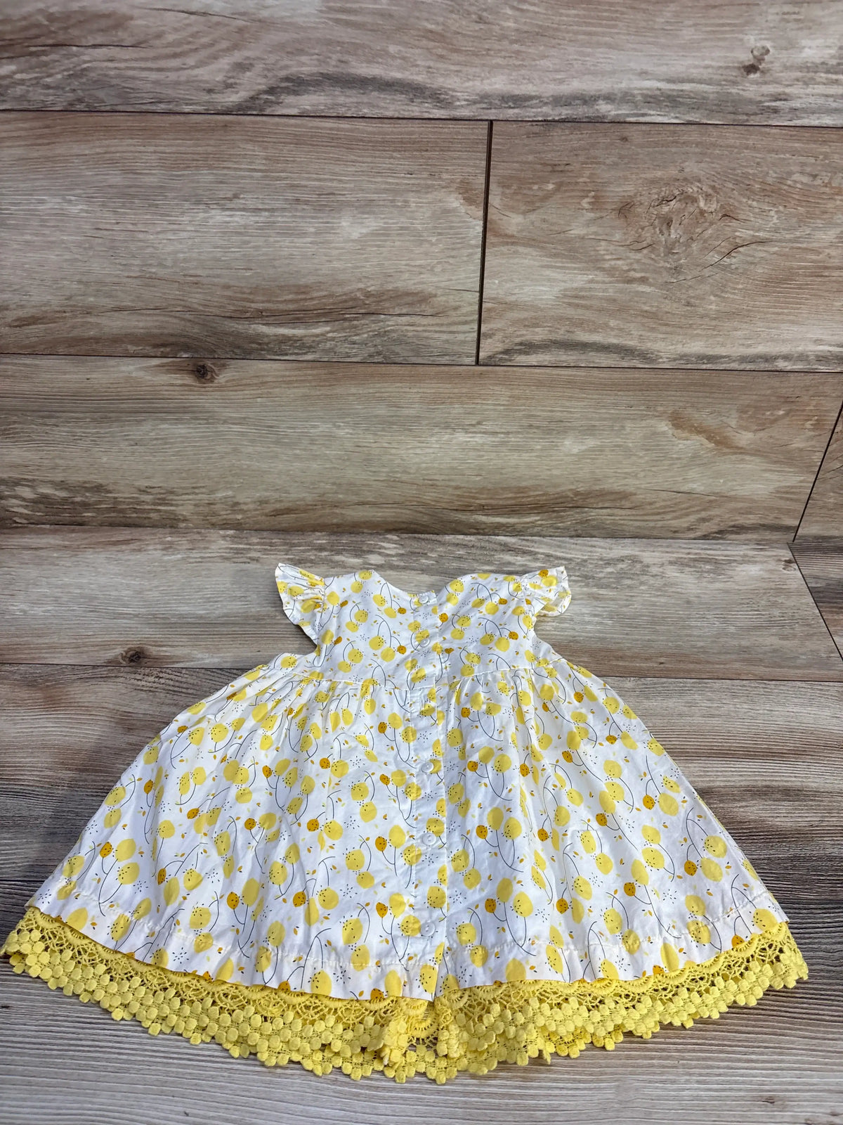 Flat lay of Du Pareil Au Meme baby lemon-print dress showcasing flutter sleeves and yellow crocheted lace hem on wood floor background