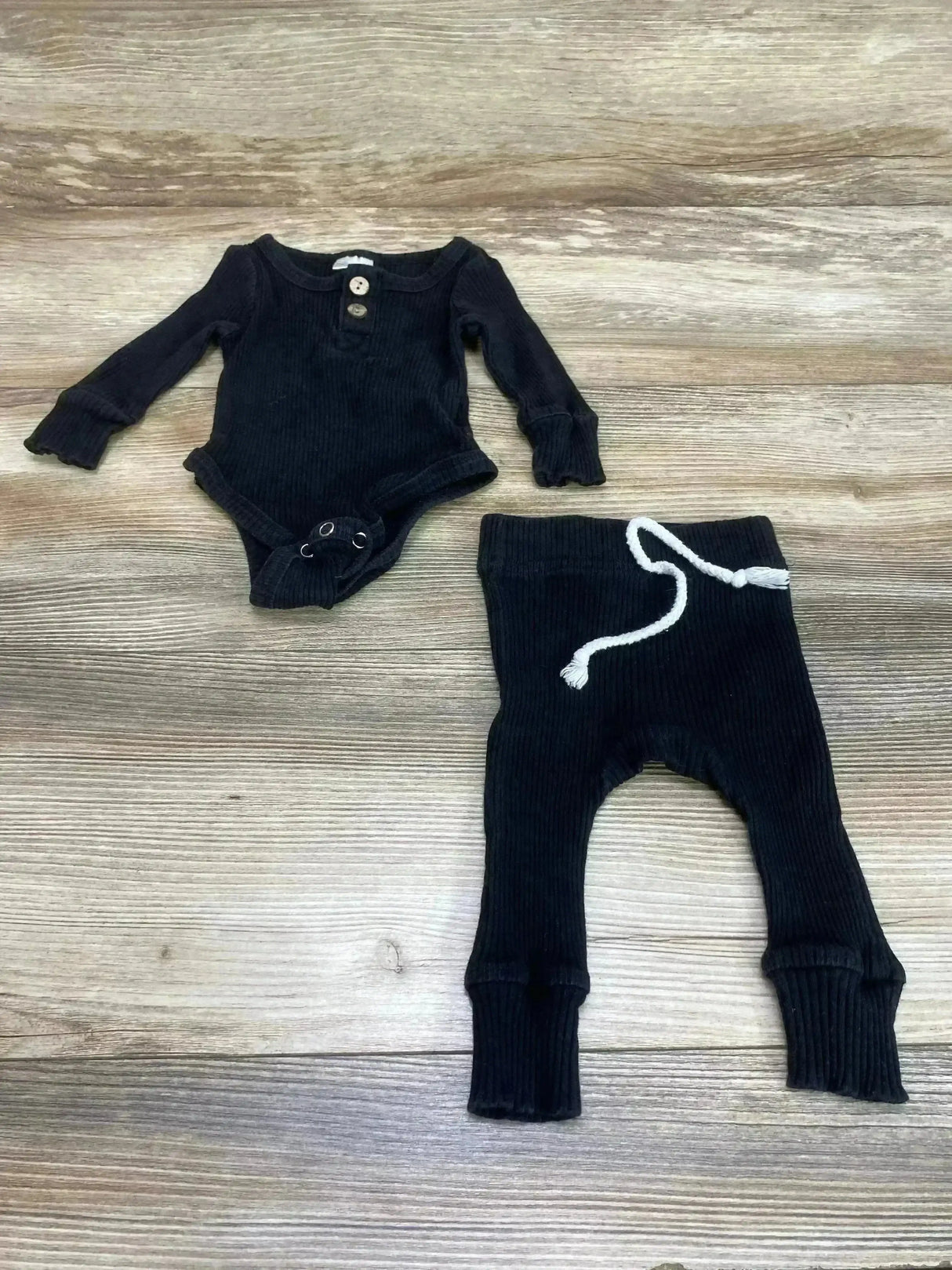 Top-down view of black ribbed baby bodysuit and matching pants showing white drawstring and wood plank background