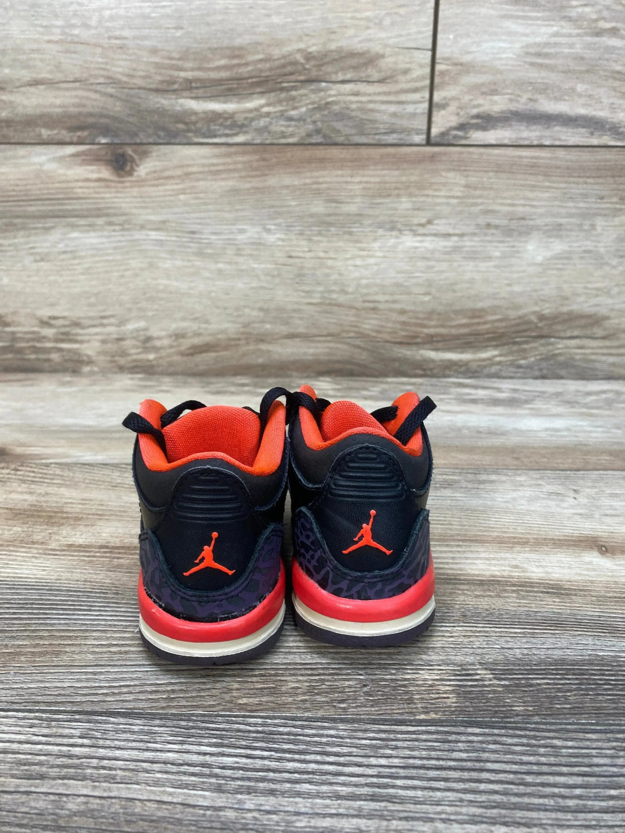 Rear view of Air Jordan 3 Retro TD toddler sneakers in black and crimson showcasing Jumpman heel logo and red inner lining on a wood plank background