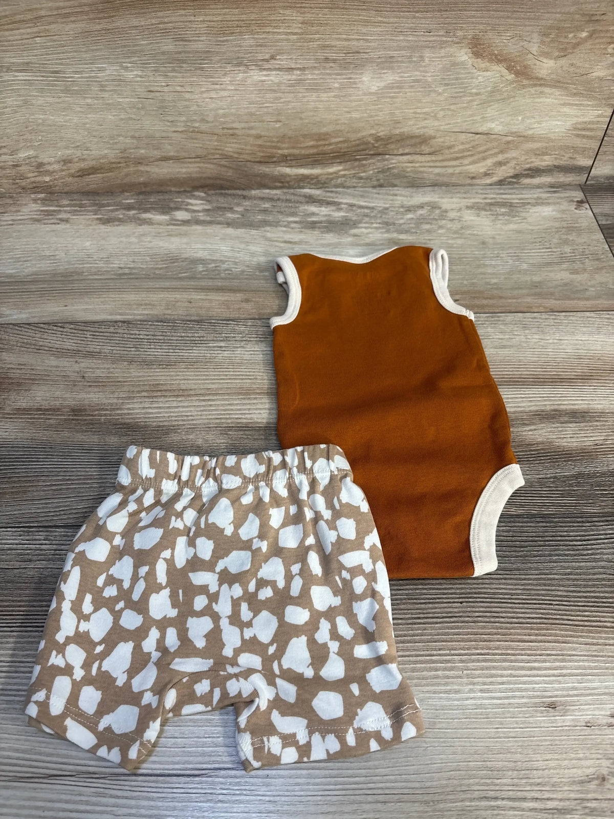 Top-down view of Okie Dokie 2pc Big Cat rust sleeveless bodysuit and tan shorts with white abstract print on wood background