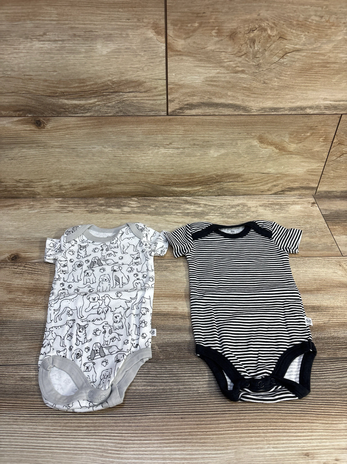 Flat lay of Honest Baby 2-pack bodysuits showing a white dog-print bodysuit with gray trim and a black-and-white striped bodysuit on wood background