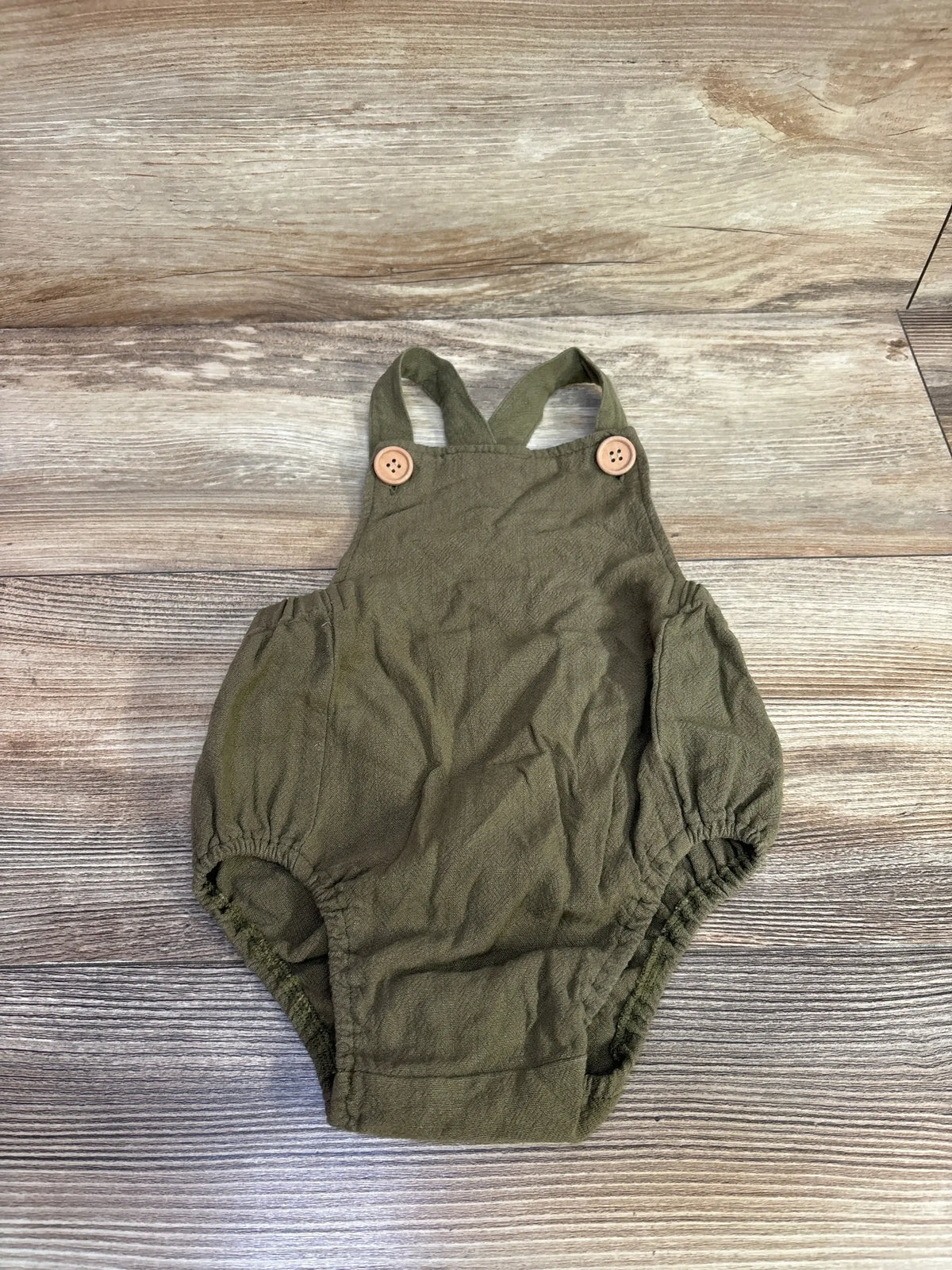 Top-down view of green linen baby romper showcasing crossed straps with wooden button closures and elasticized leg openings on wood plank background