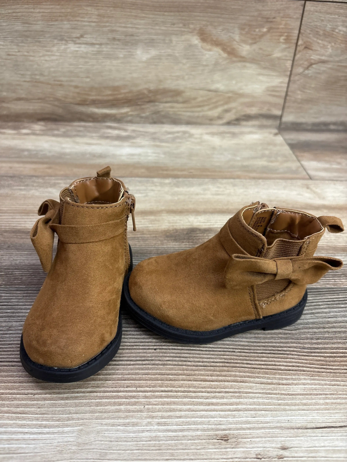 Pair of brown vegan suede baby ankle boots with decorative bows on a wooden surface.