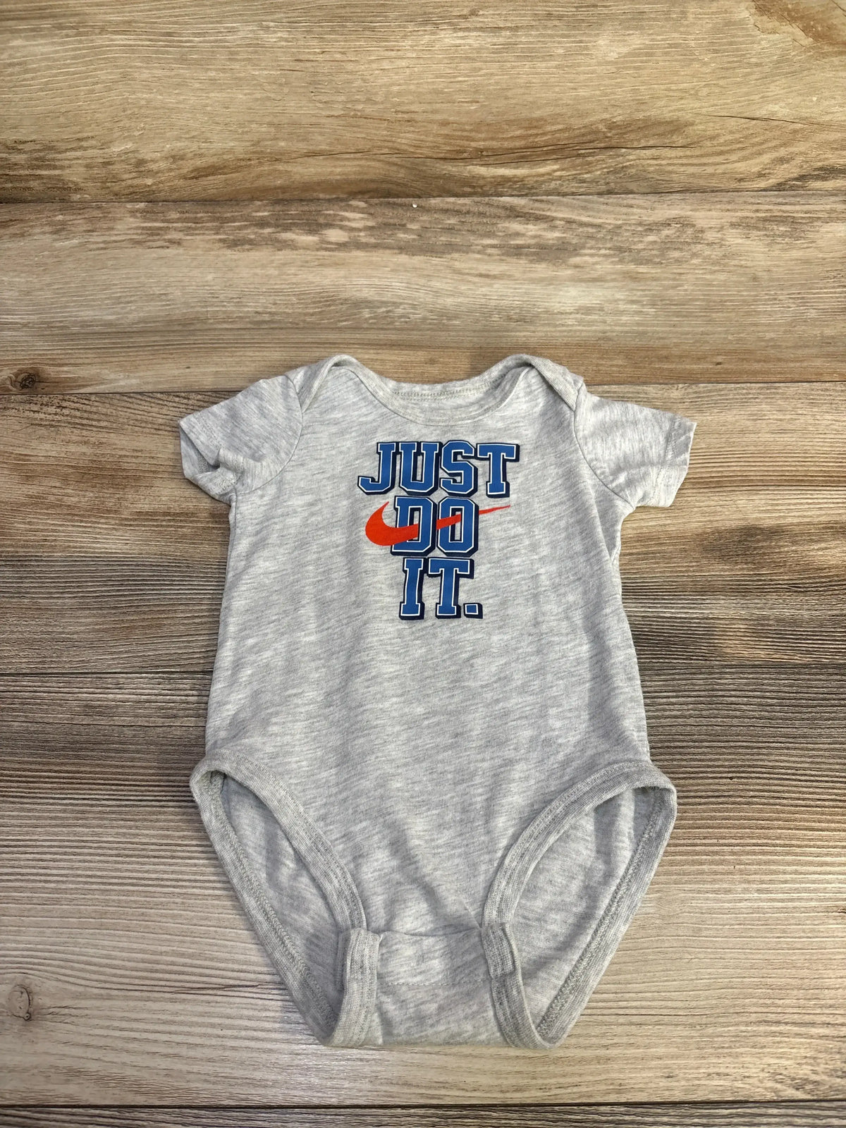 Flat lay of grey Nike 6M bodysuit showing blue JUST DO IT text with orange Nike swoosh on wood background
