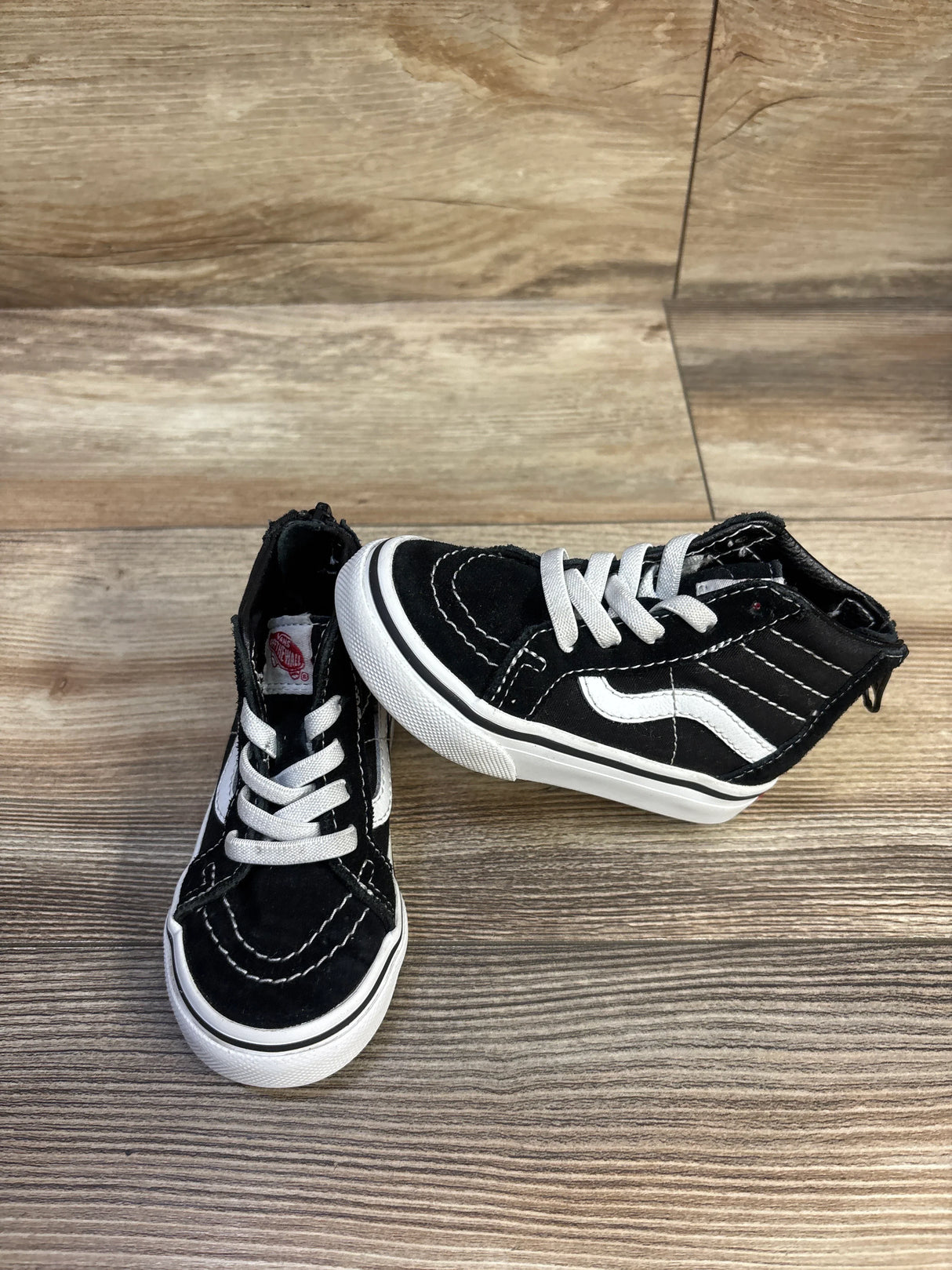 Three-quarter top view of black Vans Sk8-Hi Zip toddler sneakers showing white side stripe, white laces, and rubber sole on wood floor