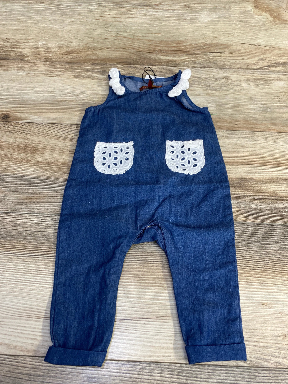 Flat lay of chambray baby jumpsuit (3-6M) showing white eyelet pockets and ruffle shoulder straps on wood background