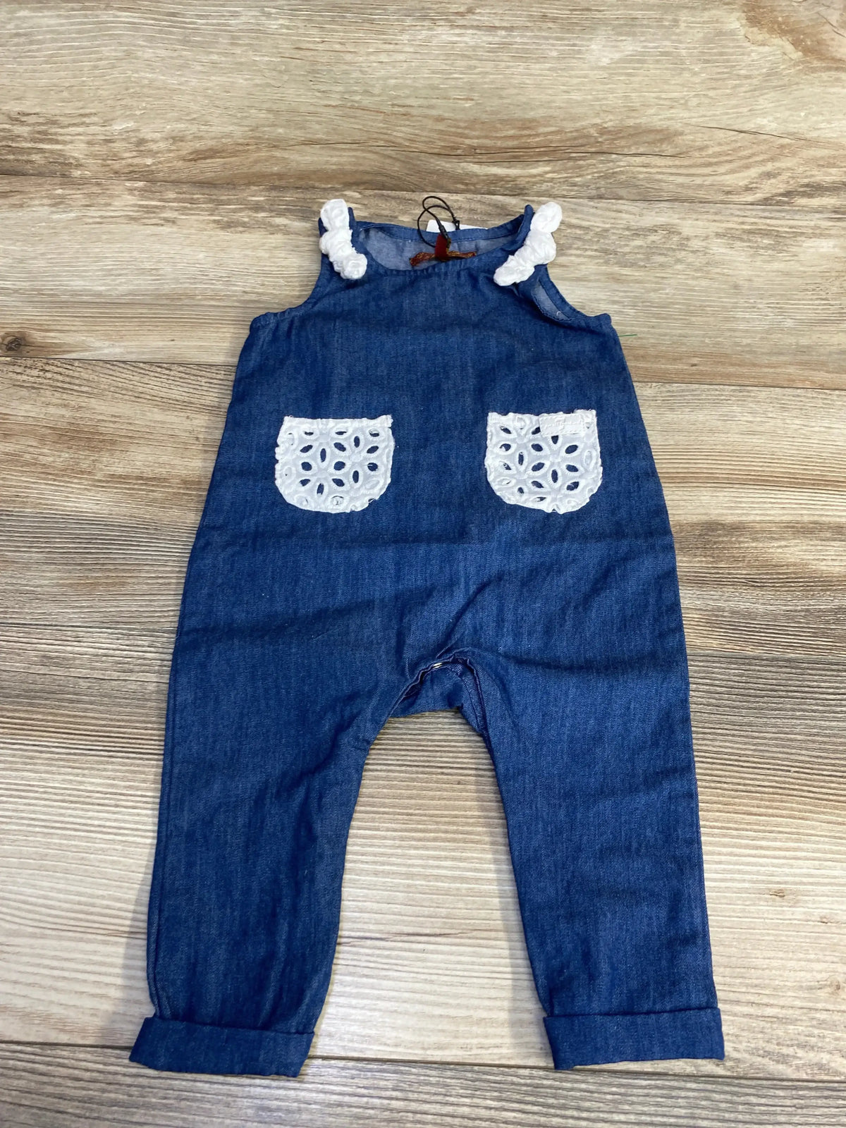 Flat lay of NEW 7 For All Mankind Chambray Jumpsuit size 3-6m showcasing blue chambray sleeveless jumpsuit with white eyelet pockets on wood plank surface