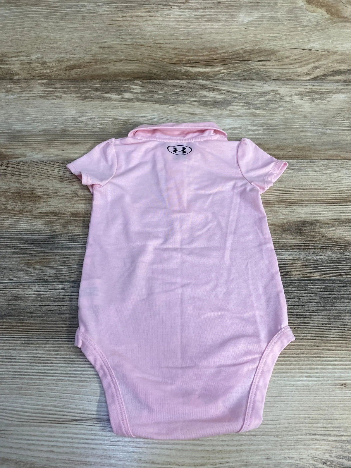 Back view of Under Armour Polo Bodysuit Pink size 6-9m showcasing light pink short sleeve bodysuit with collar and brand logo on wood plank floor